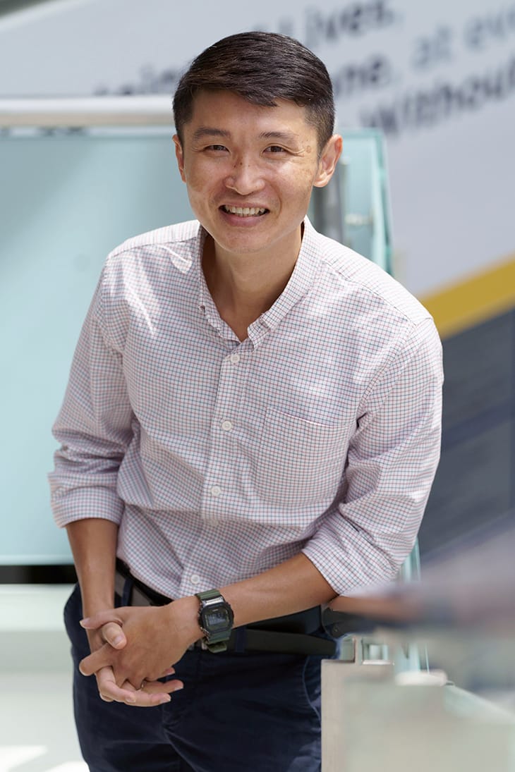 Asian man in a plaid shirt with a Casio G-Shock watch, standing and smiling.
