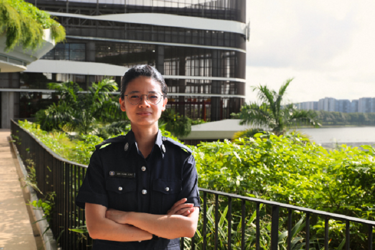 Officer in dark uniform with arms crossed; foliage, buildings, water in background.