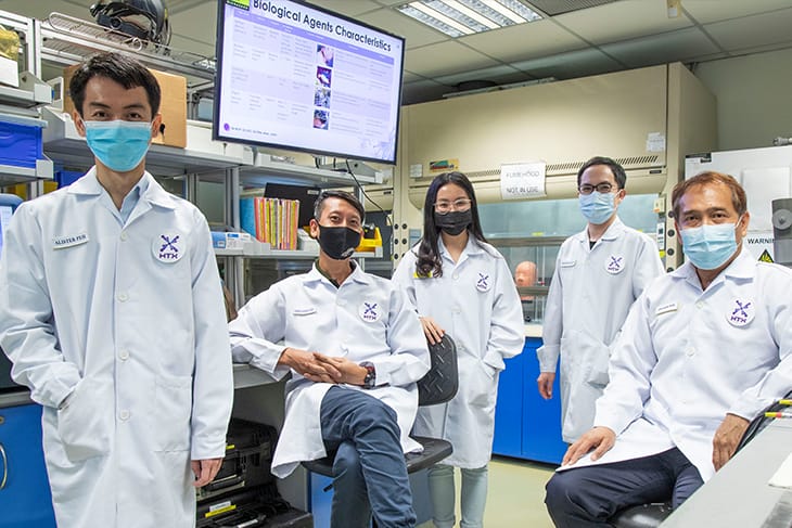 Five HTX employees in lab coats and masks pose in a lab with "Biological Agents" chart on a screen.