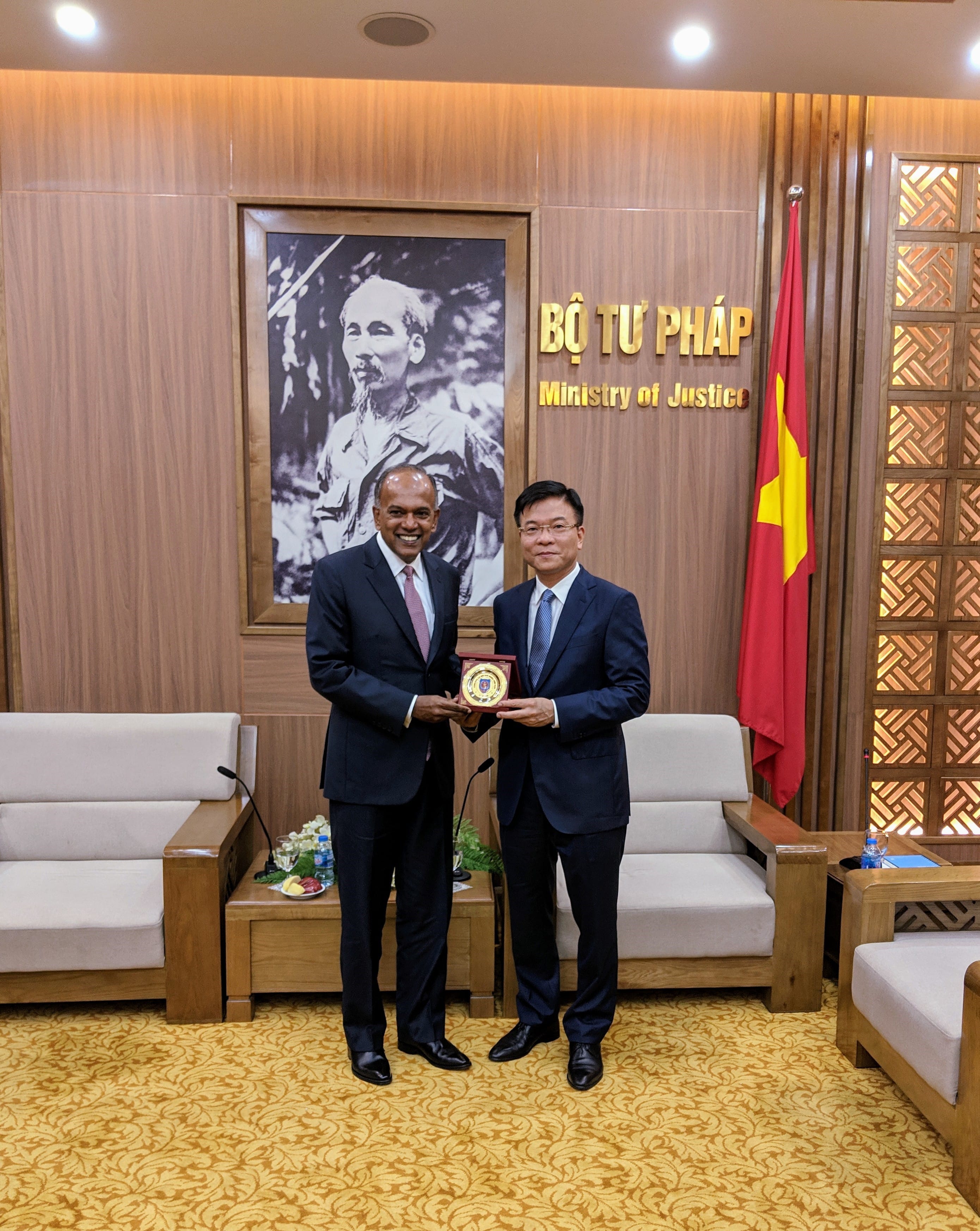 Two men in suits present a golden plaque, Vietnam flag, Ho Chi Minh portrait in background.