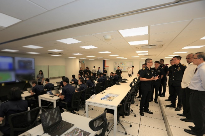 Control room with people in uniform, others observe, and monitors displaying data.