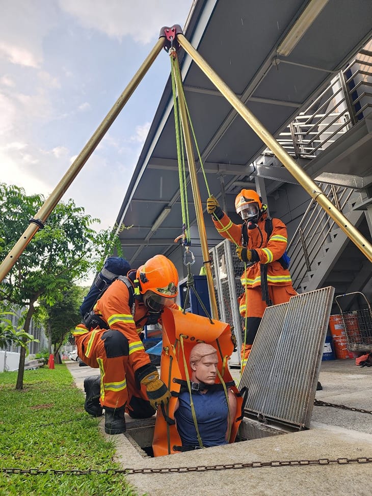 Two firefighters hoist a mannequin from a manhole using a golden tripod and ropes. They wear orange gear and helmets.