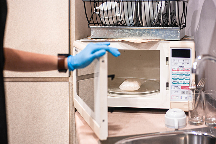 Gloved hand opens microwave; wrapped item on turntable. Dish rack on top, sink nearby.