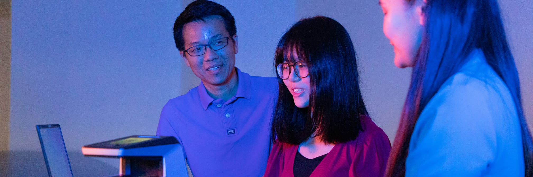 Three people, with glasses, stand near a computer in blue and red lighting.