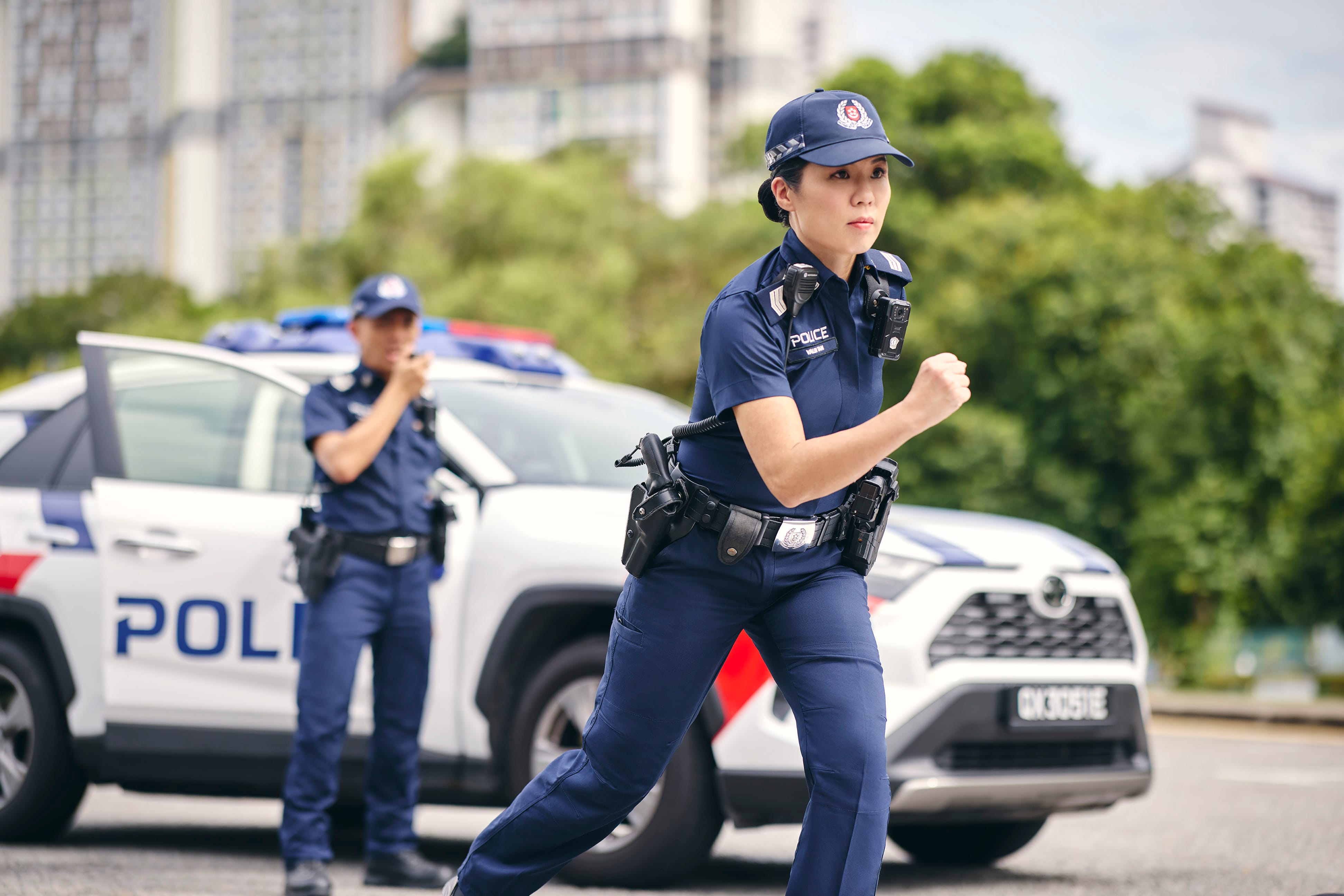 Female police officer running towards an incident while another officer communicates via radio beside a patrol car.