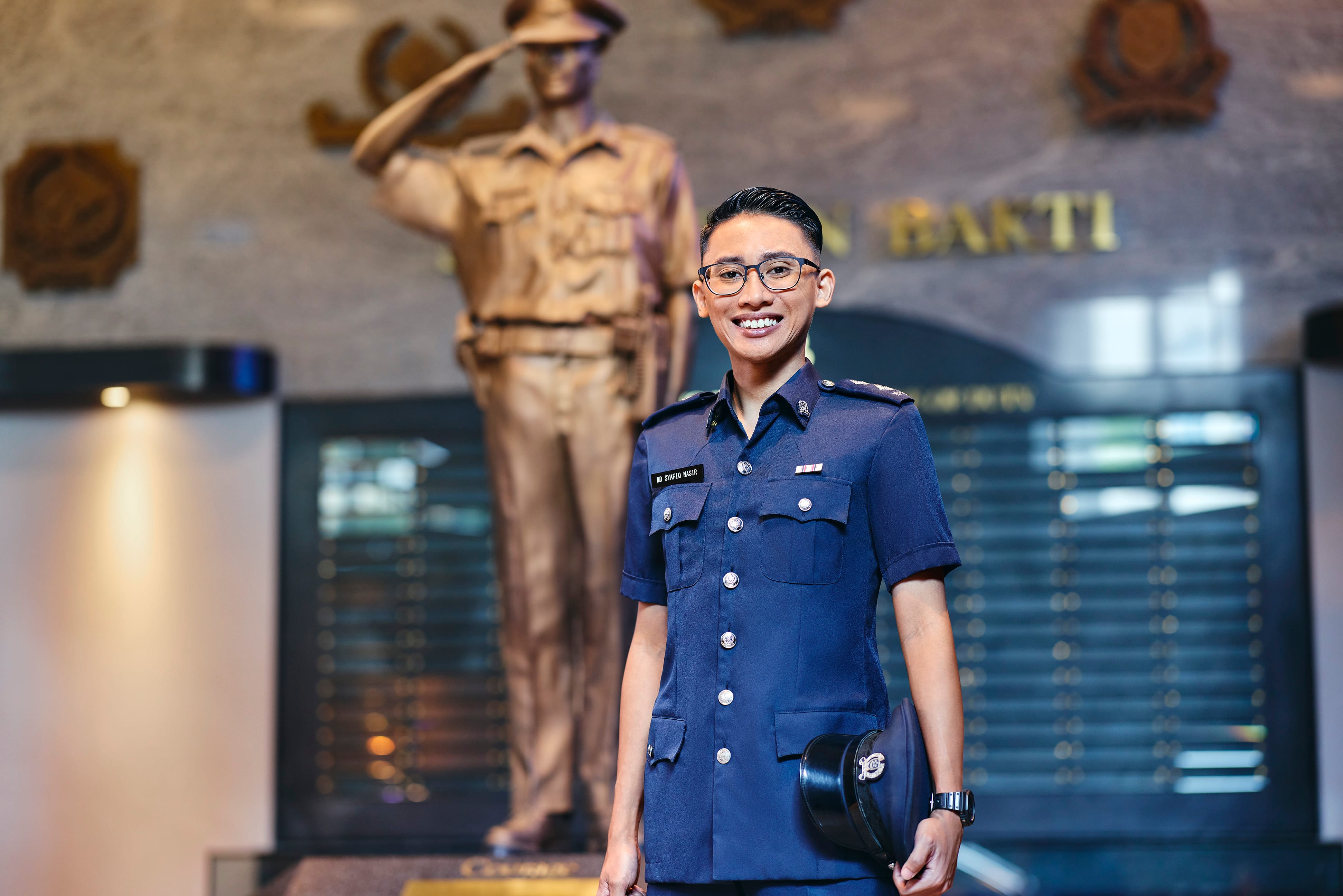 Police officer in full uniform smiling confidently while standing in front of a statue of a saluting officer.