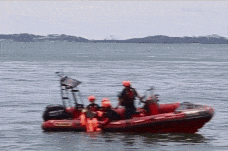 Four people in a red inflatable boat on water, mountains in background.