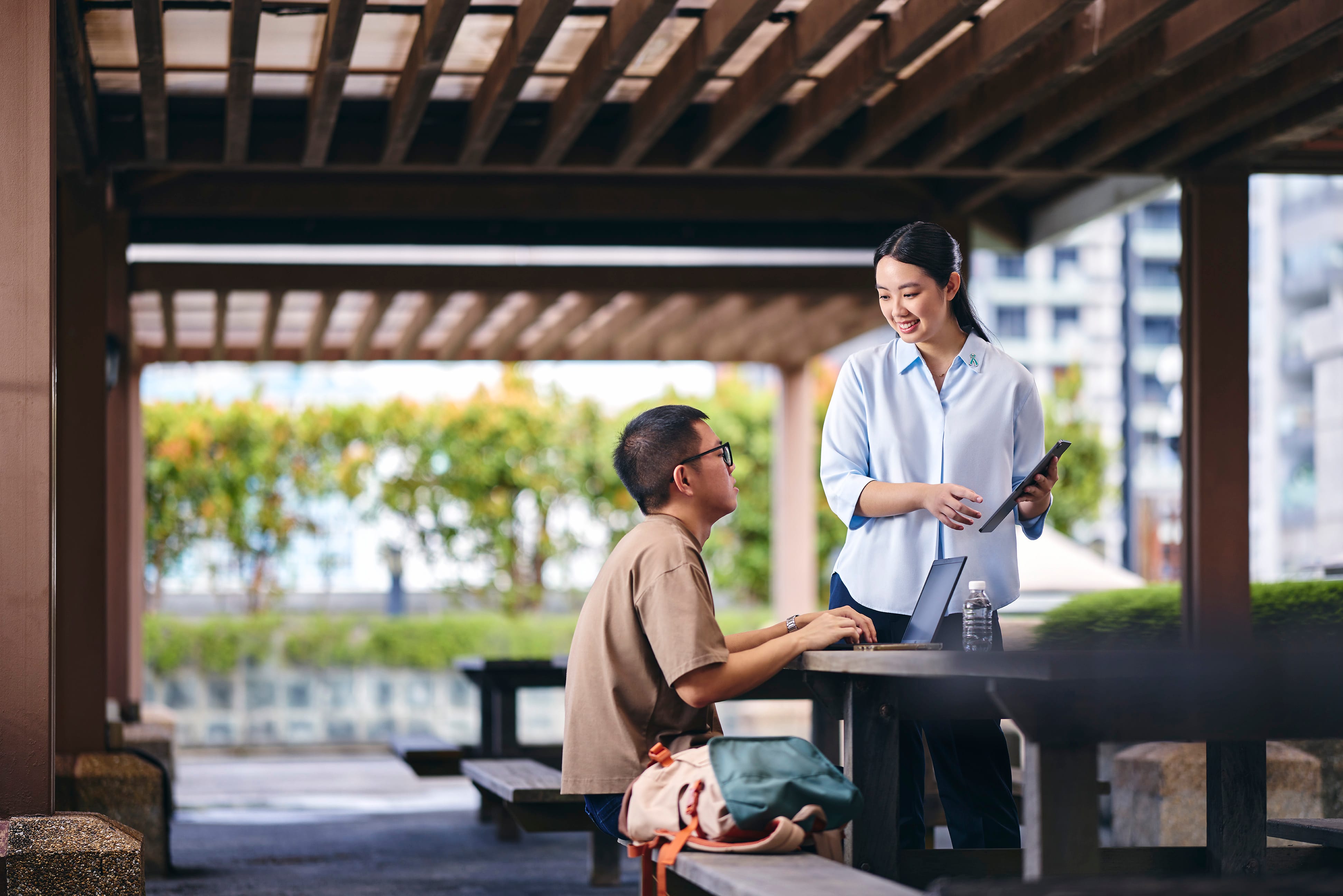 Woman in a light blue blouse holding a tablet and talking with a man seated at a table outdoors.