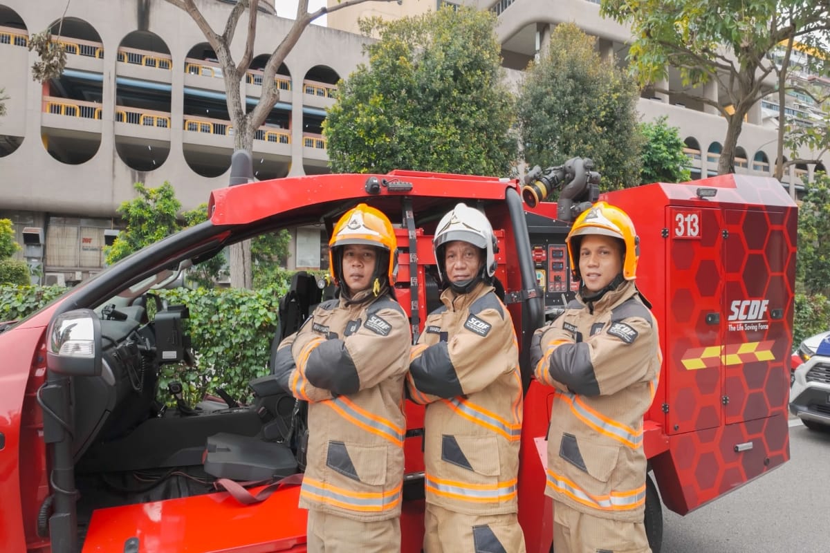 Three SCDF firefighters in uniform stand by a red fire rescue vehicle.