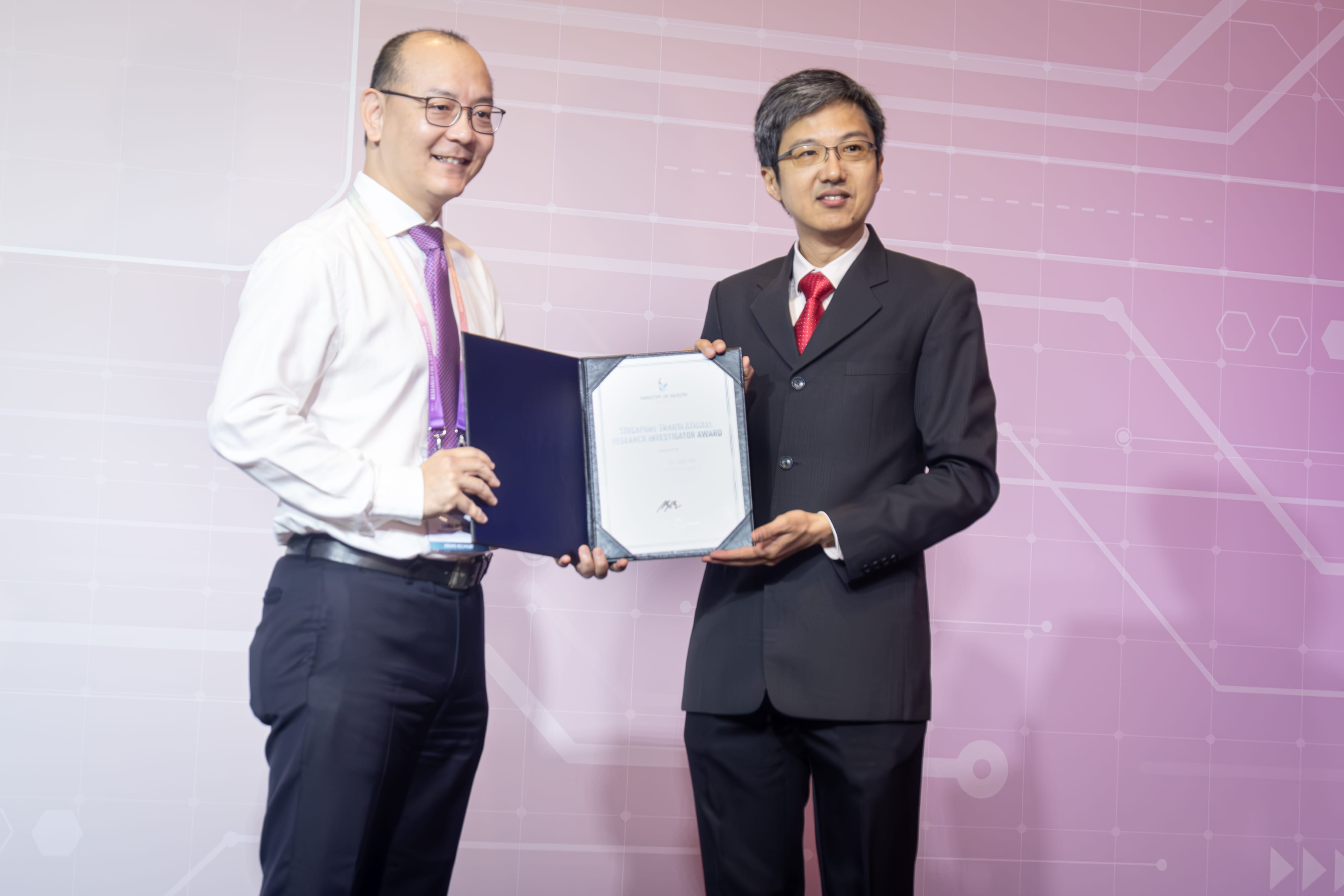 Two individuals stand, one holding a framed certificate, against a purple and white background with geometric patterns.