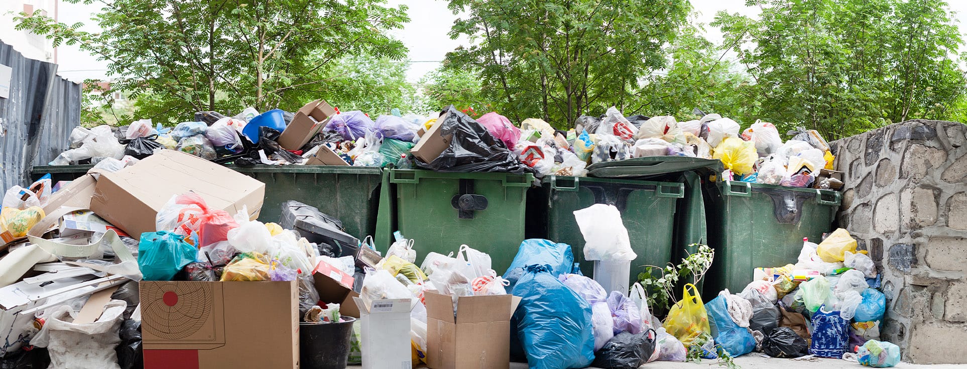 Overflowing garbage bins with bags and boxes of trash, surrounded by trees and a stone wall.