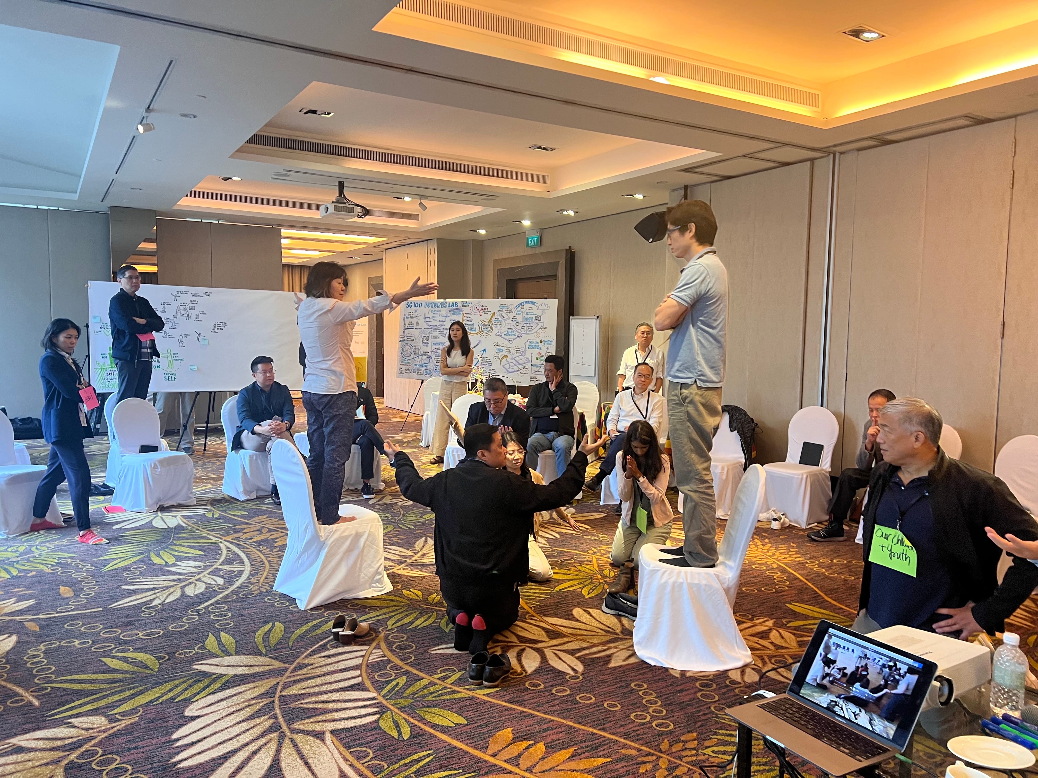 A group of people in a conference room are engaged in an interactive activity with chairs and presentation boards.