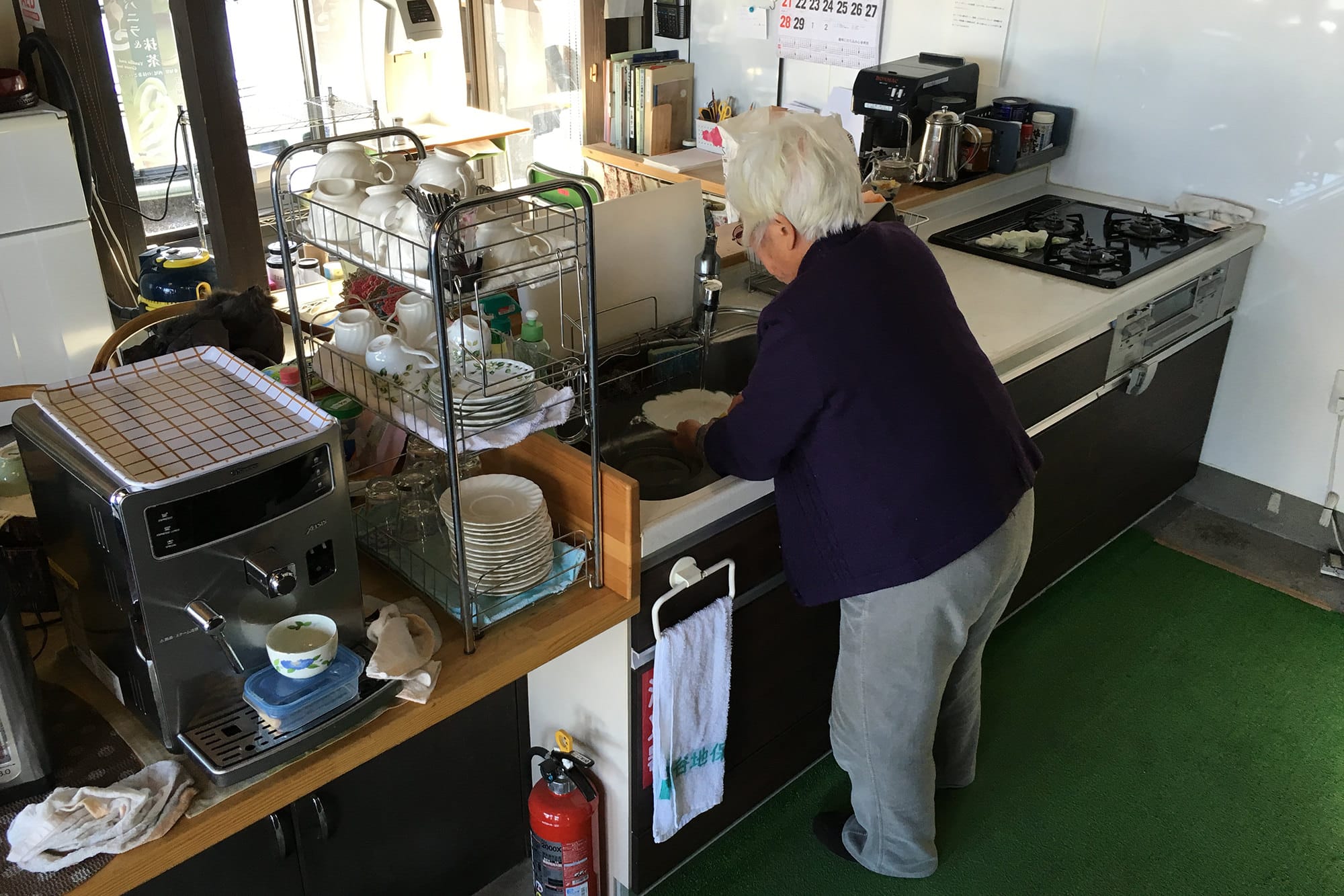 An elderly person with white hair washes dishes in a cluttered kitchen with various appliances and utensils.