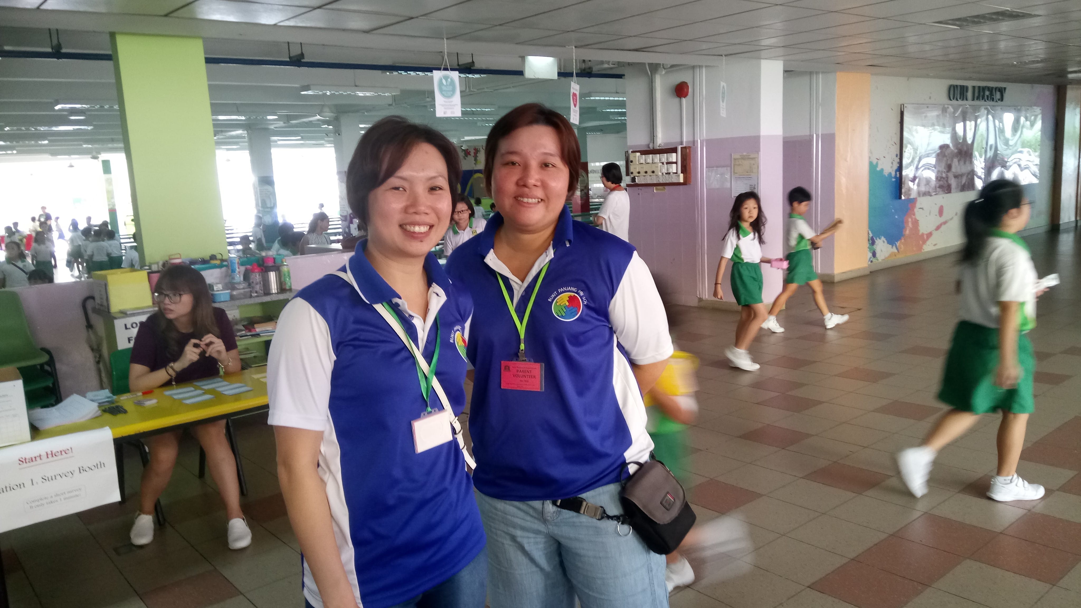 Two people in blue shirts with name tags stand indoors near a yellow table; children walk in the background.