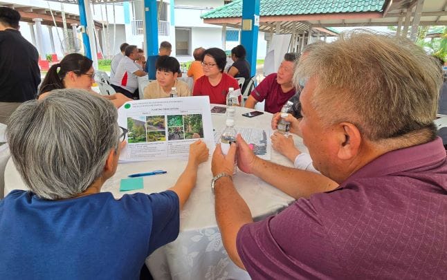 Two elderly people discuss a document with photos at a table in a communal area with others in the background.
