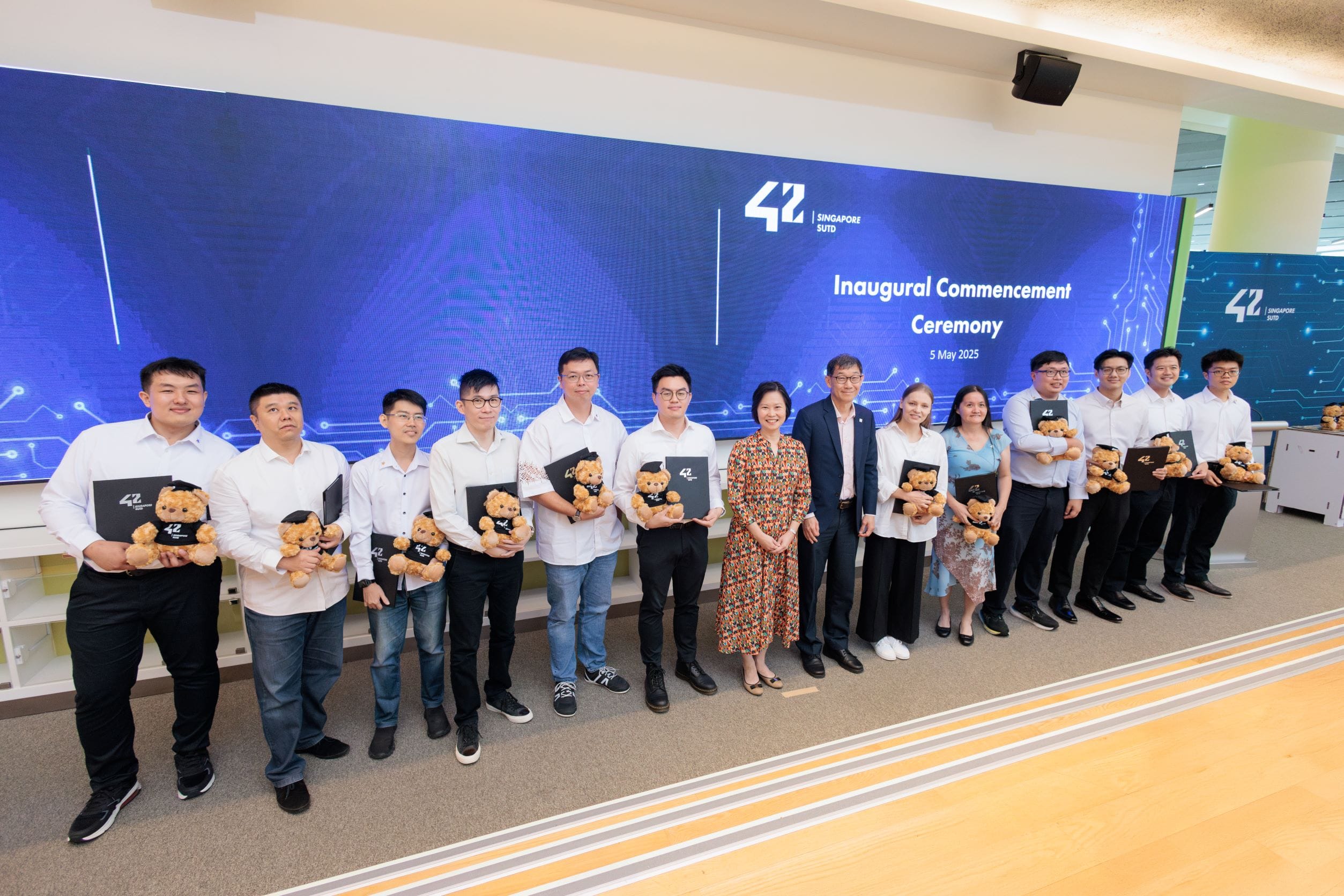 Group of people standing in front of a screen at an inaugural commencement ceremony, some holding certificates and teddy bears.