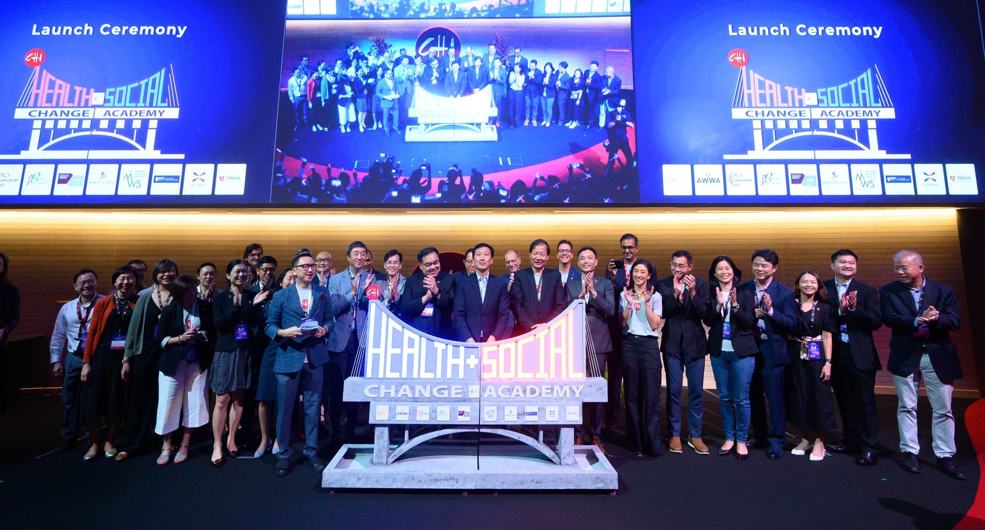 Group of people standing and clapping on a stage with a health and social change academy banner and a launch ceremony sign above.