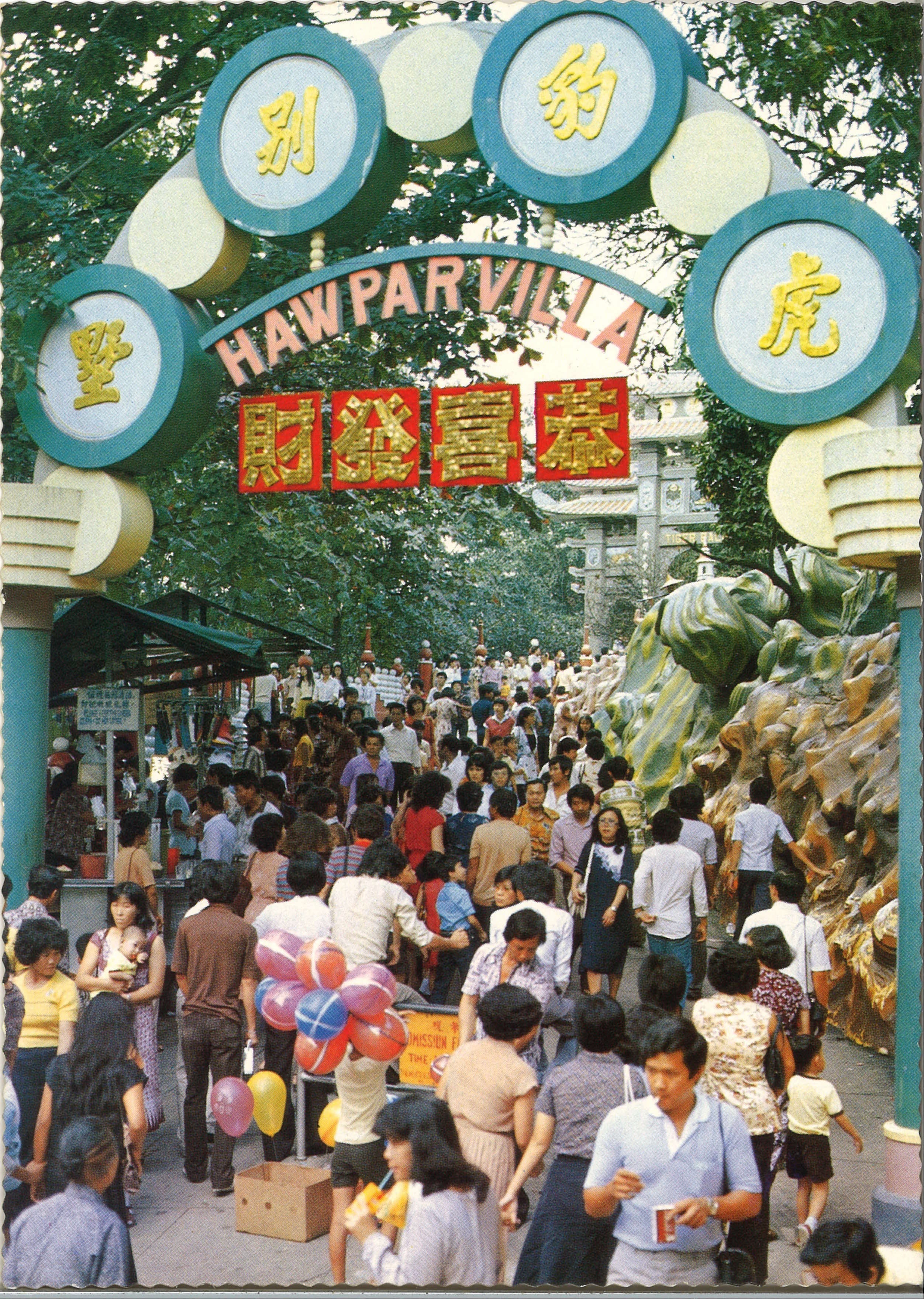 Crowded entrance of Haw Par Villa, with colorful balloons and people walking under decorated archway in front of rock formations.