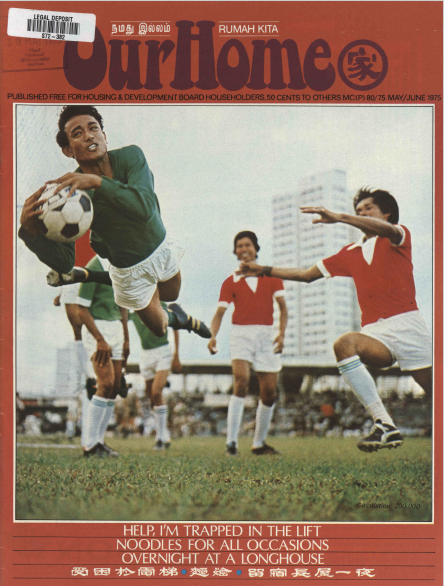 A soccer player in green jumps and kicks a ball while players in red jerseys approach and tall buildings stand in the background.
