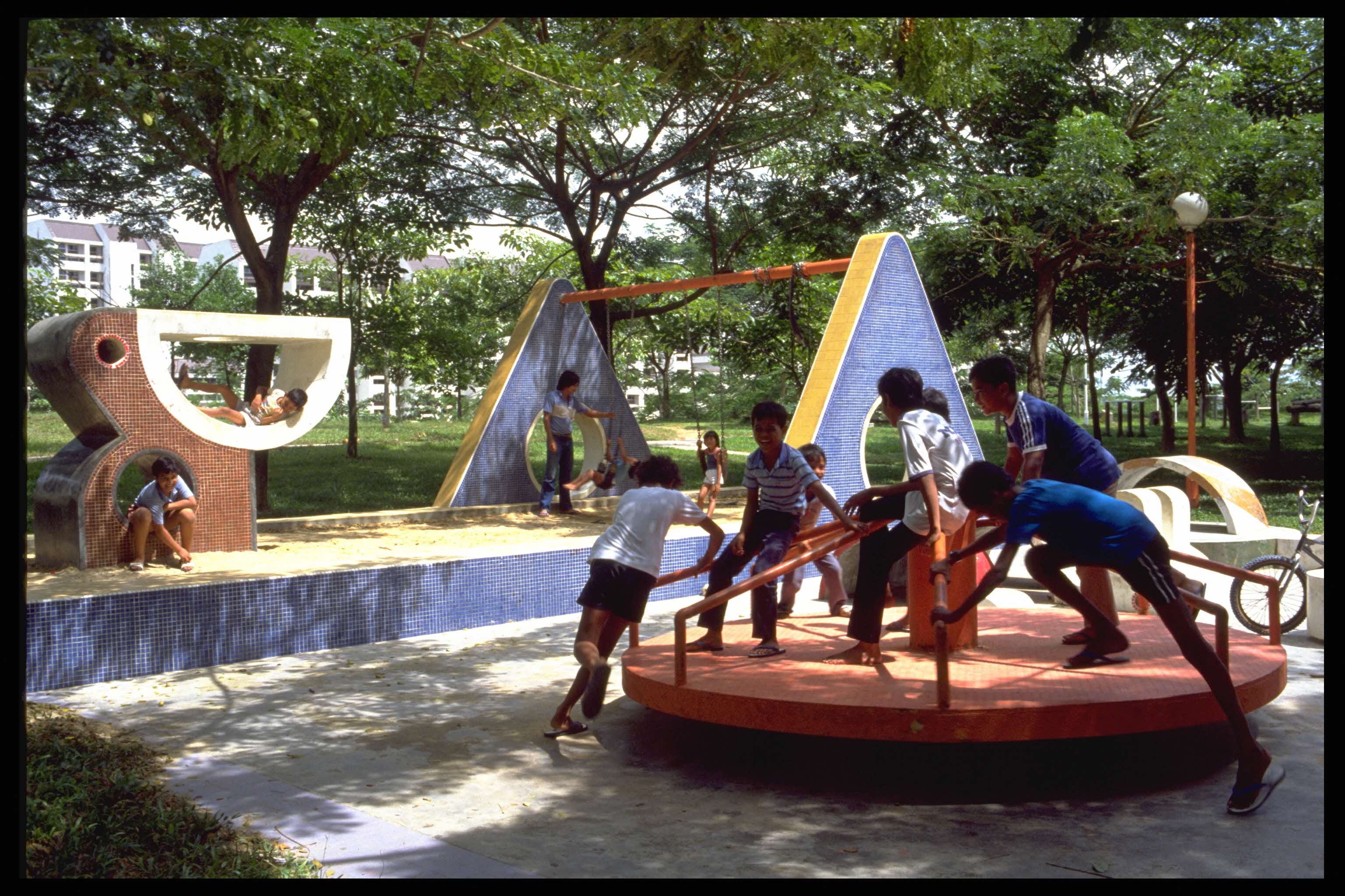 Children playing on various playground equipment surrounded by trees in an outdoor park.