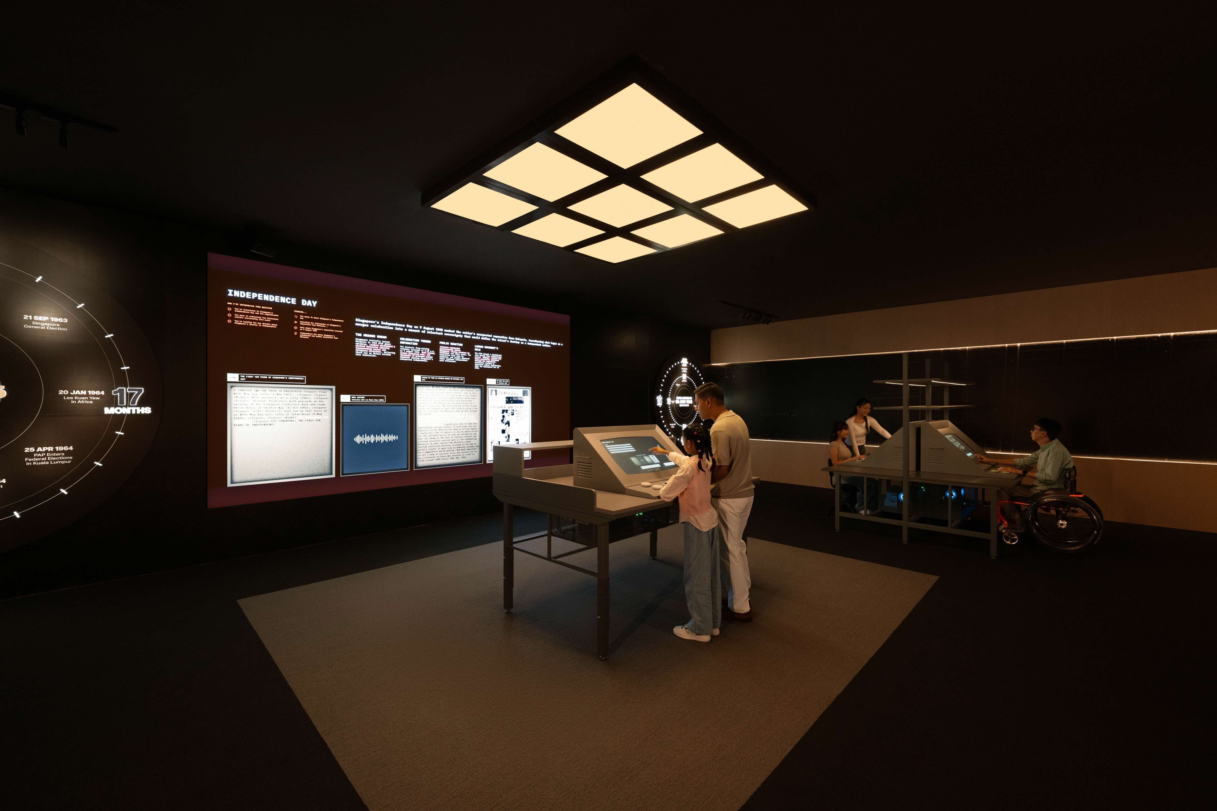 People interact with touch screens in a dark room featuring a large Independence Day display and circular timeline on the wall.