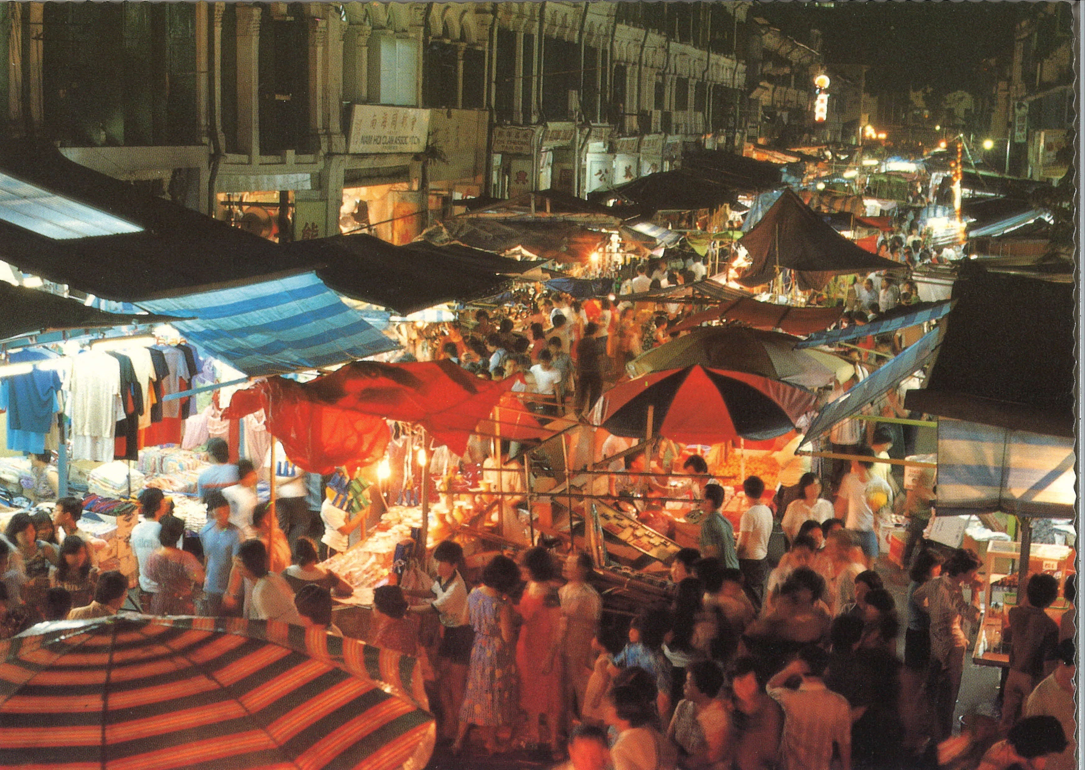 A crowded night market with various stalls and umbrellas, selling clothes and goods under colorful lights.