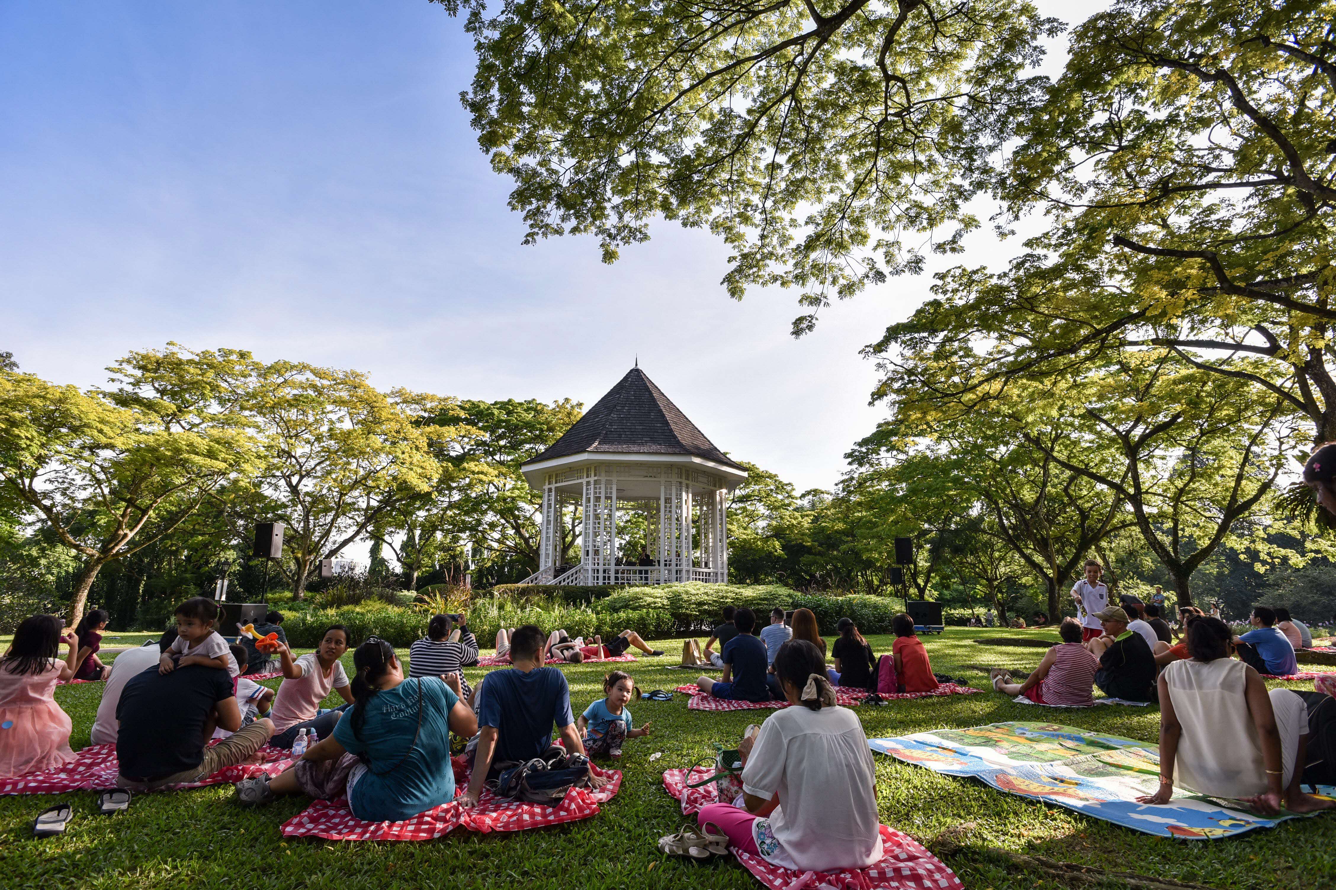 People are sitting on blankets on grass, facing a white bandstand surrounded by green trees under a blue sky.