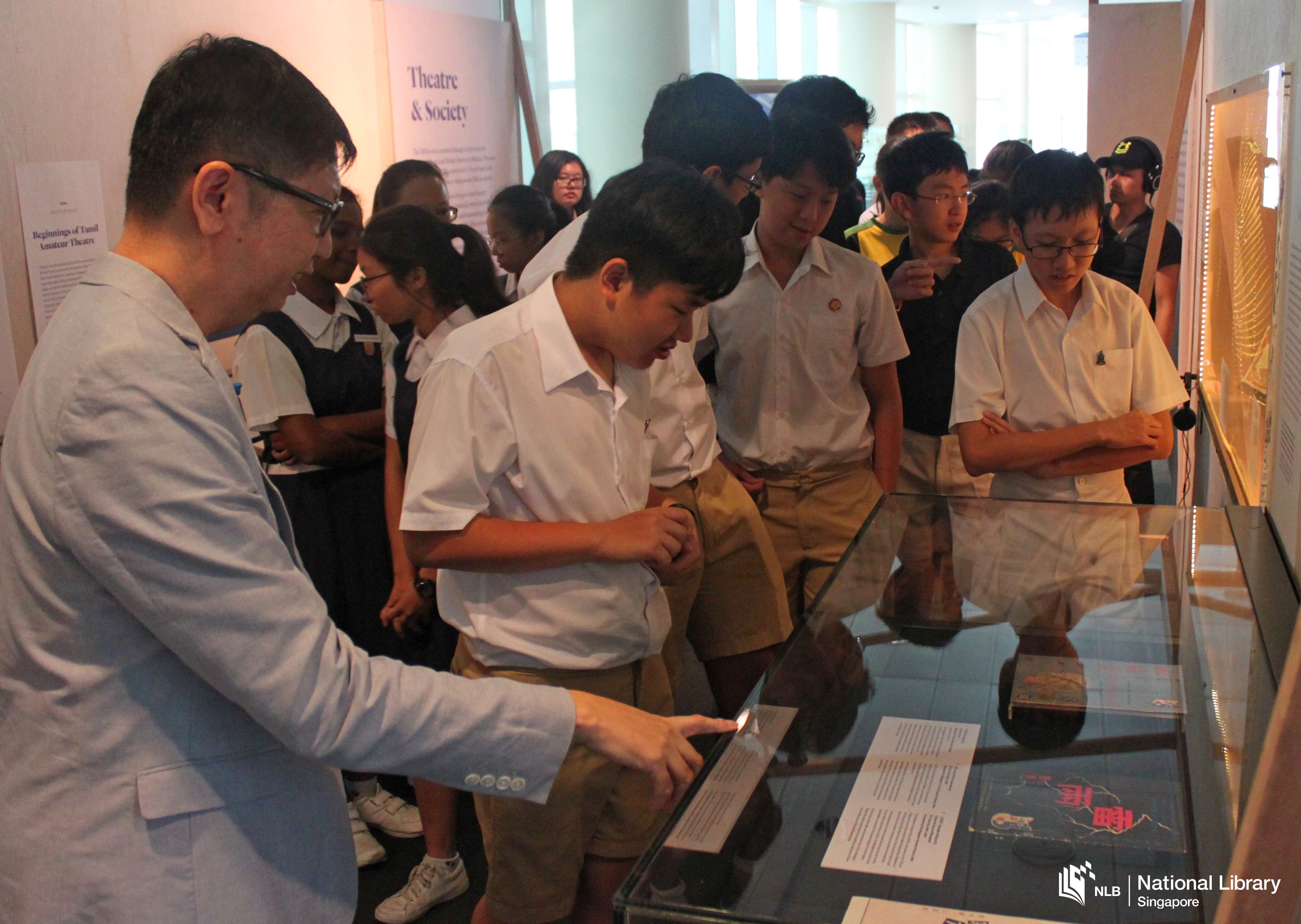 A group of students in uniforms observing a display in a museum or library, with a person explaining the exhibit.