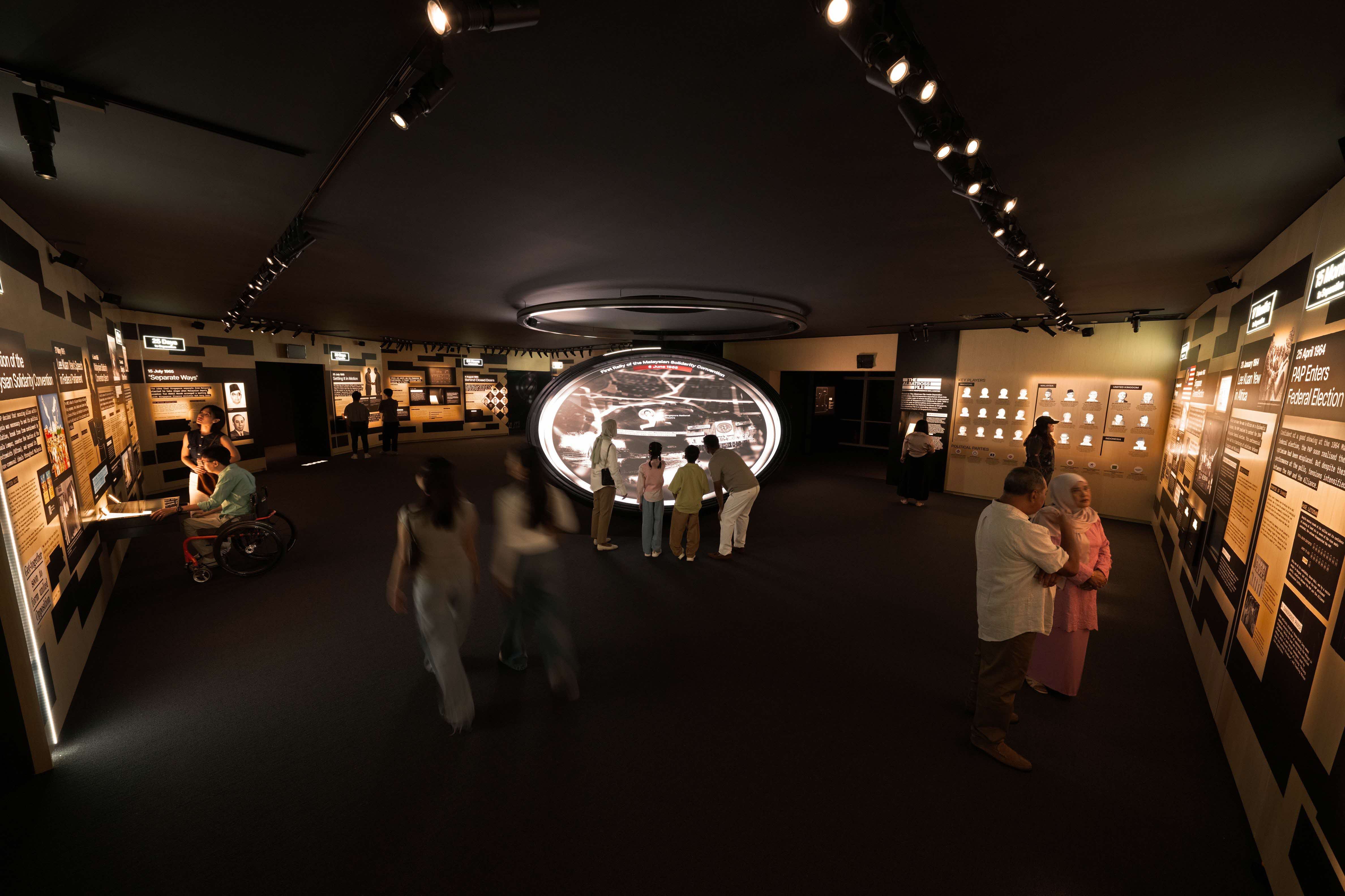 Visitors inside a dimly lit museum interact with exhibits and read informational panels along the walls.
