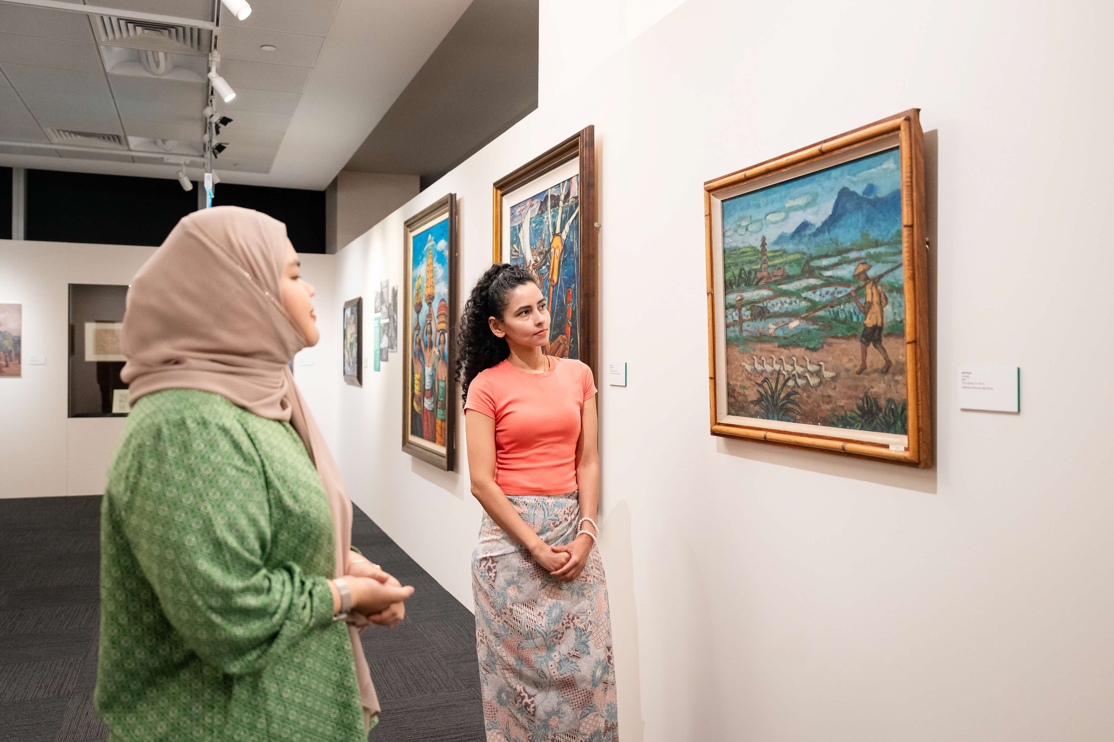 Two women in a gallery observe colorful paintings depicting various scenes.