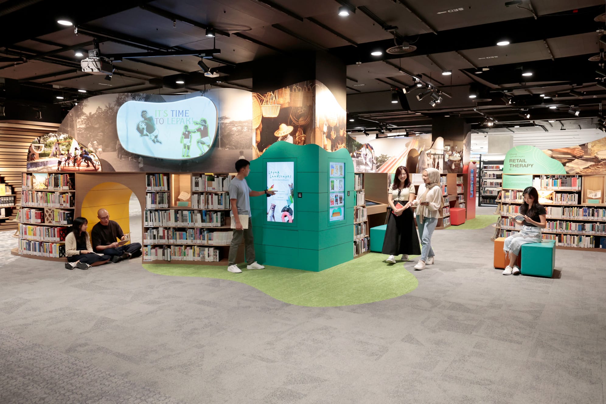 Library interior with bookshelves, people reading and interacting around a digital kiosk, and a screen displaying "It's Time to Lepak."