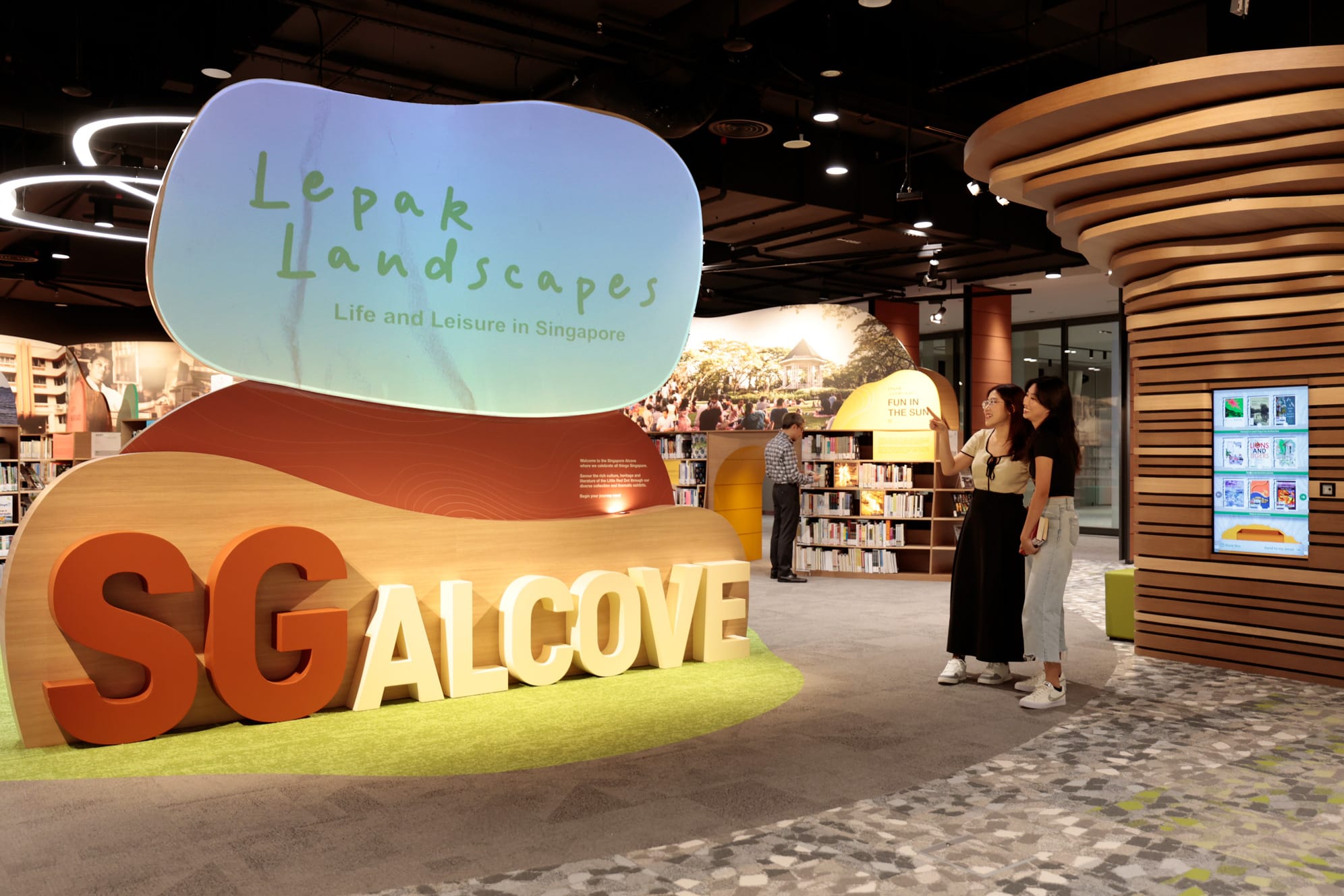Exhibit with "Lepak Landscapes" and "SG ALcove" signs, bookshelves, and a digital display in a modern library setting.