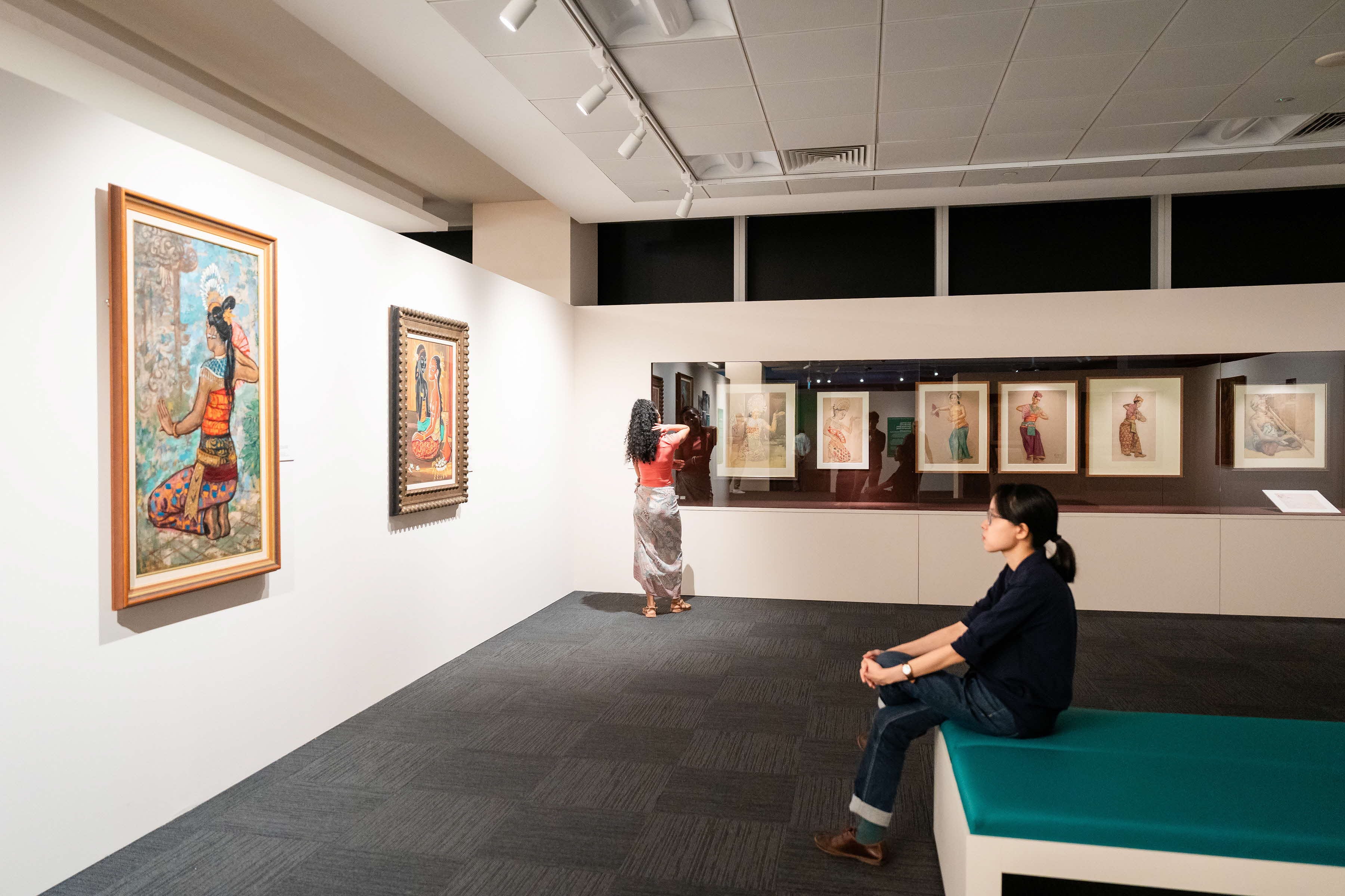 A person looks at art pieces on a museum wall while another sits on a bench in the foreground.