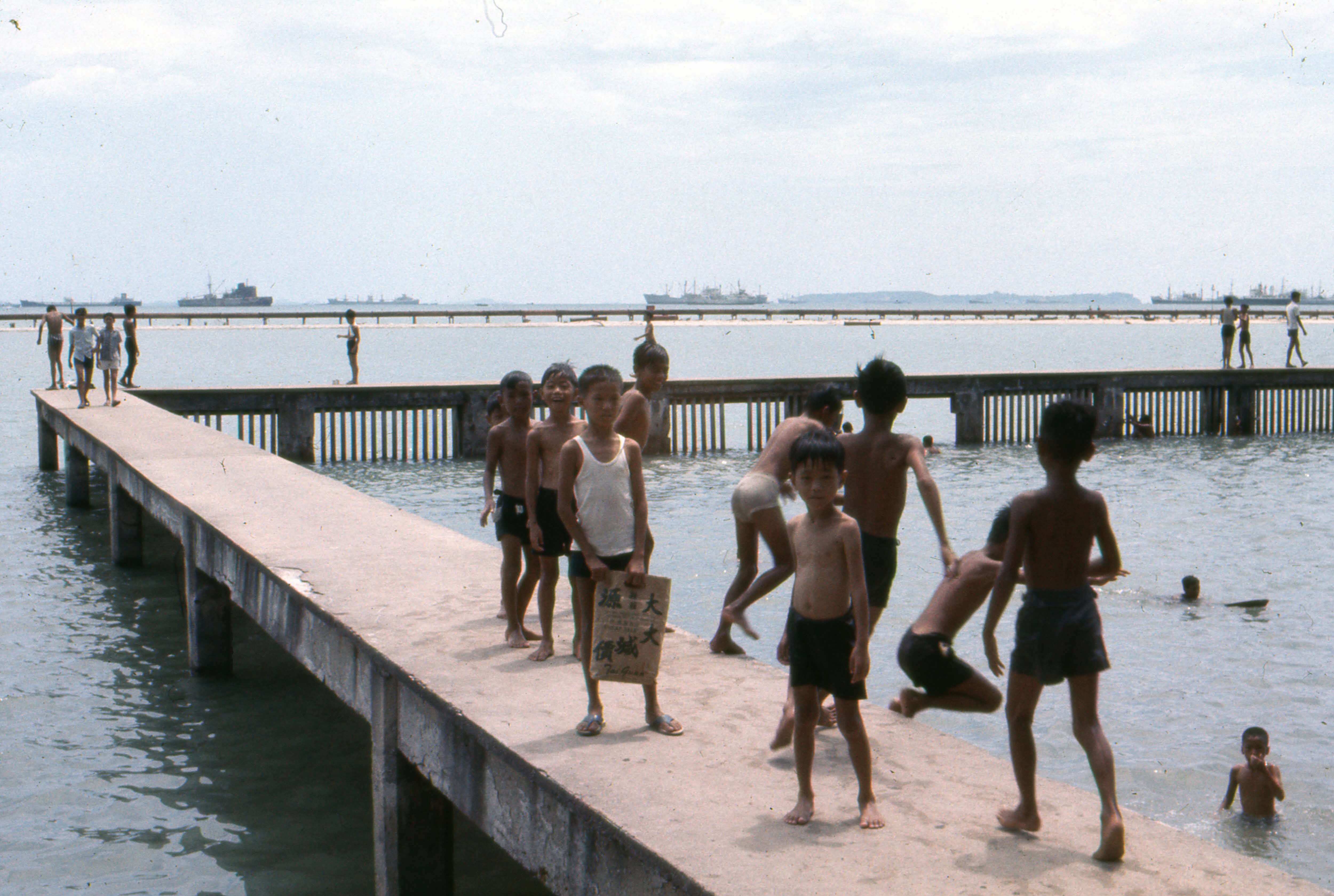 Children in swimwear are playing and walking on an elevated concrete platform over water, with distant ships visible.