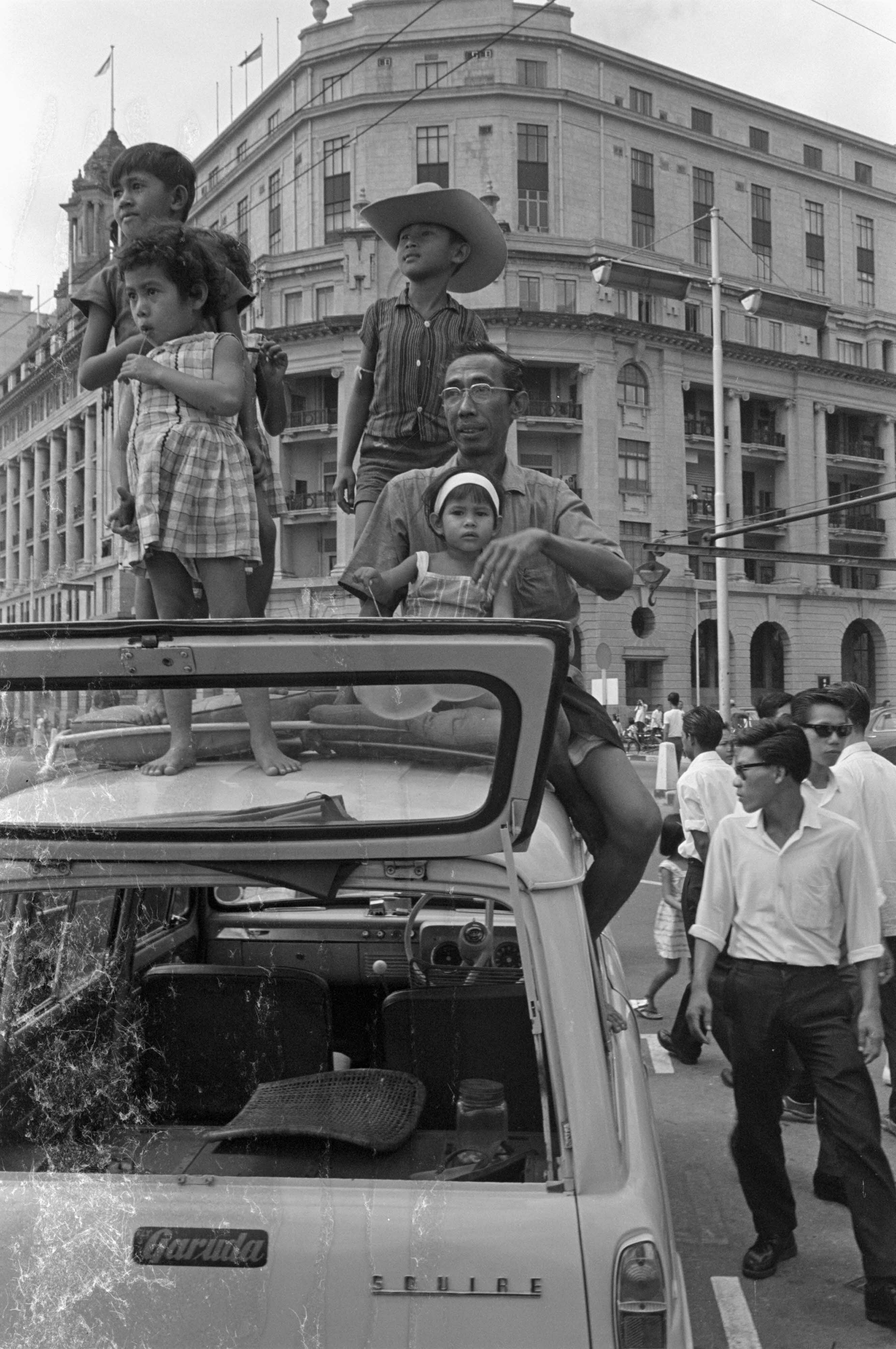 Several people stand on top of a parked car in front of a large, ornate building in a busy street scene.