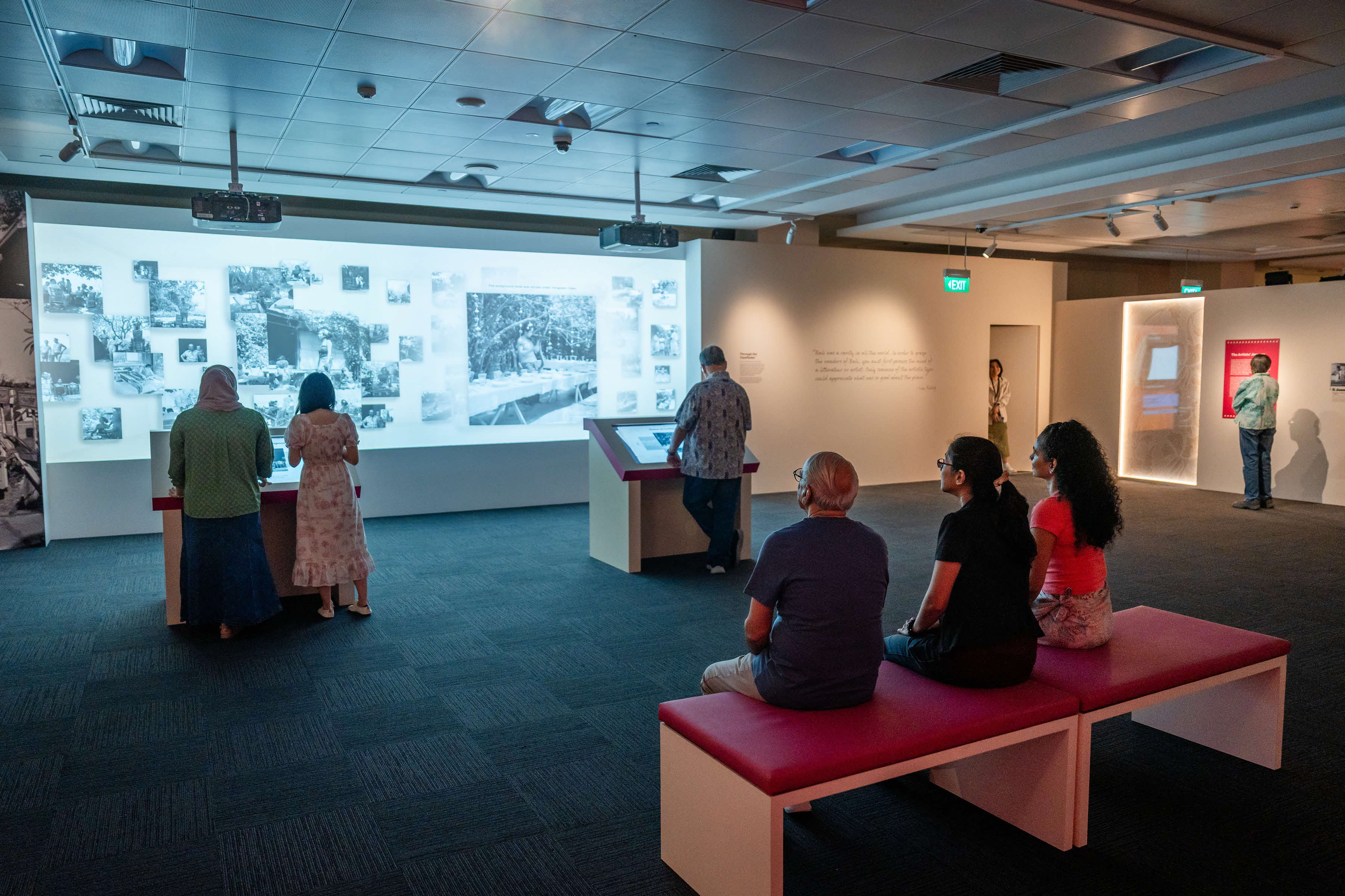 People observing displays in an exhibit room, with two sitting on a bench and others standing near interactive screens.