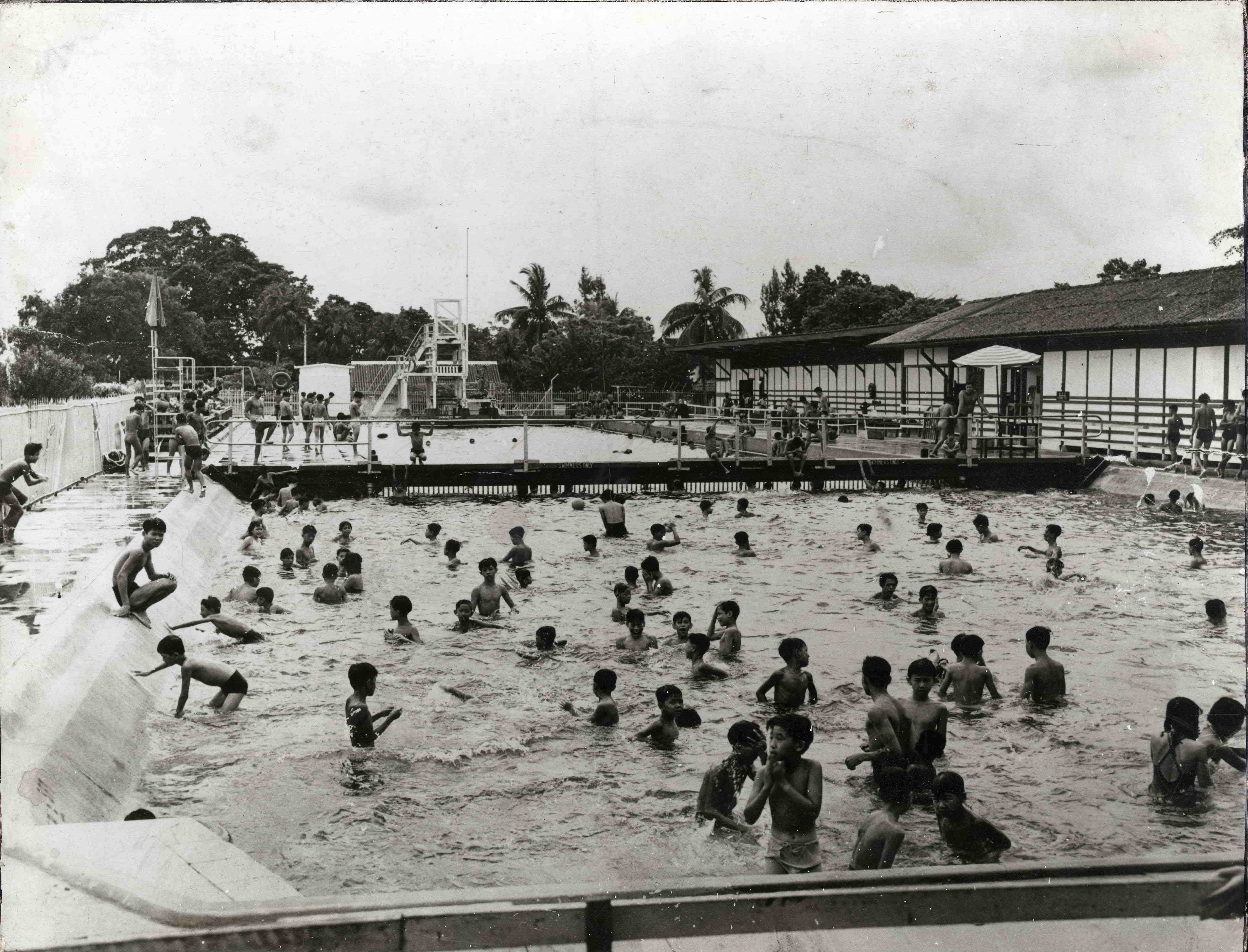 A crowded outdoor swimming pool with many people swimming and buildings in the background surrounded by trees.