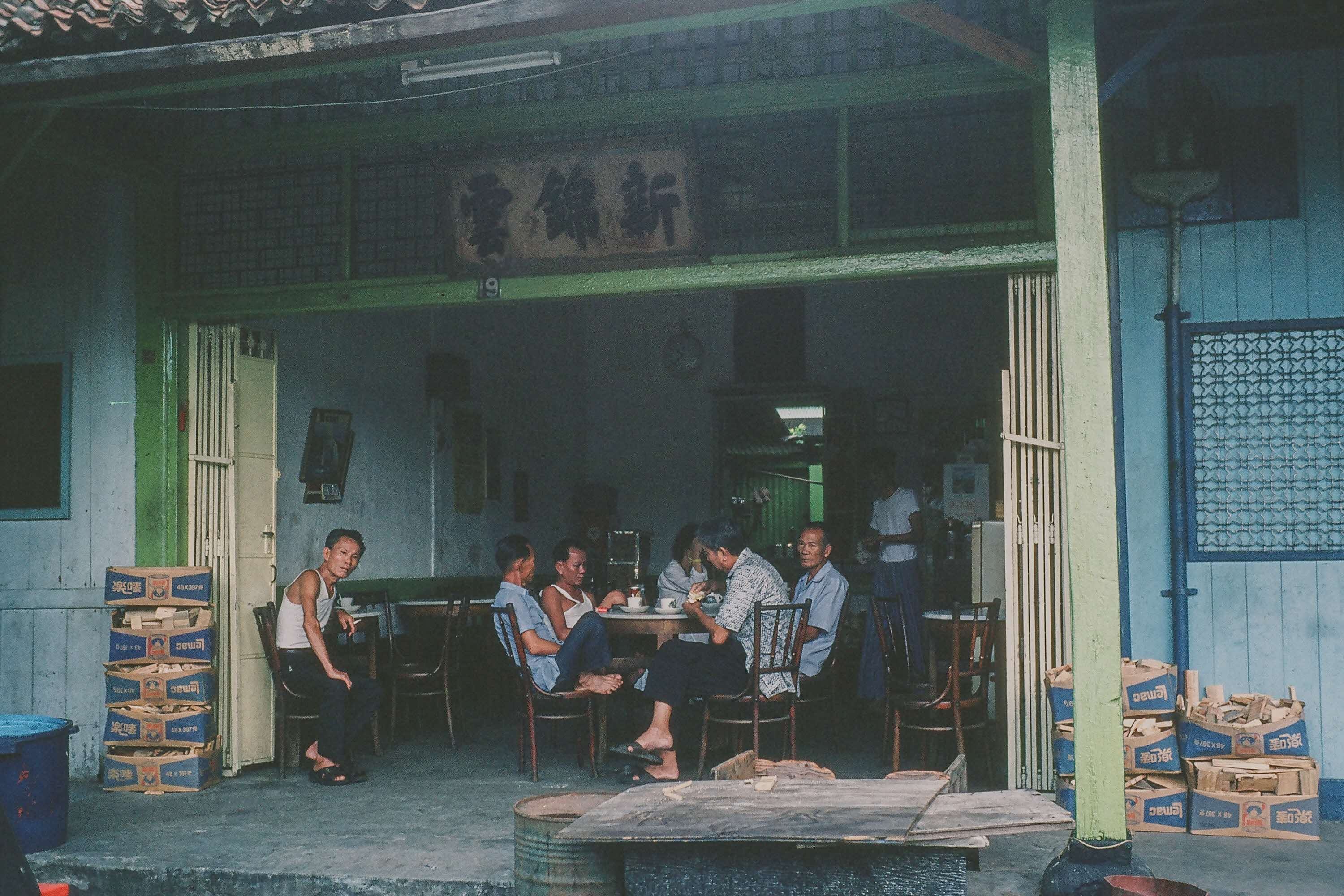 Several people sitting and conversing around tables in an open-air building, with stacked boxes and green-painted doors.