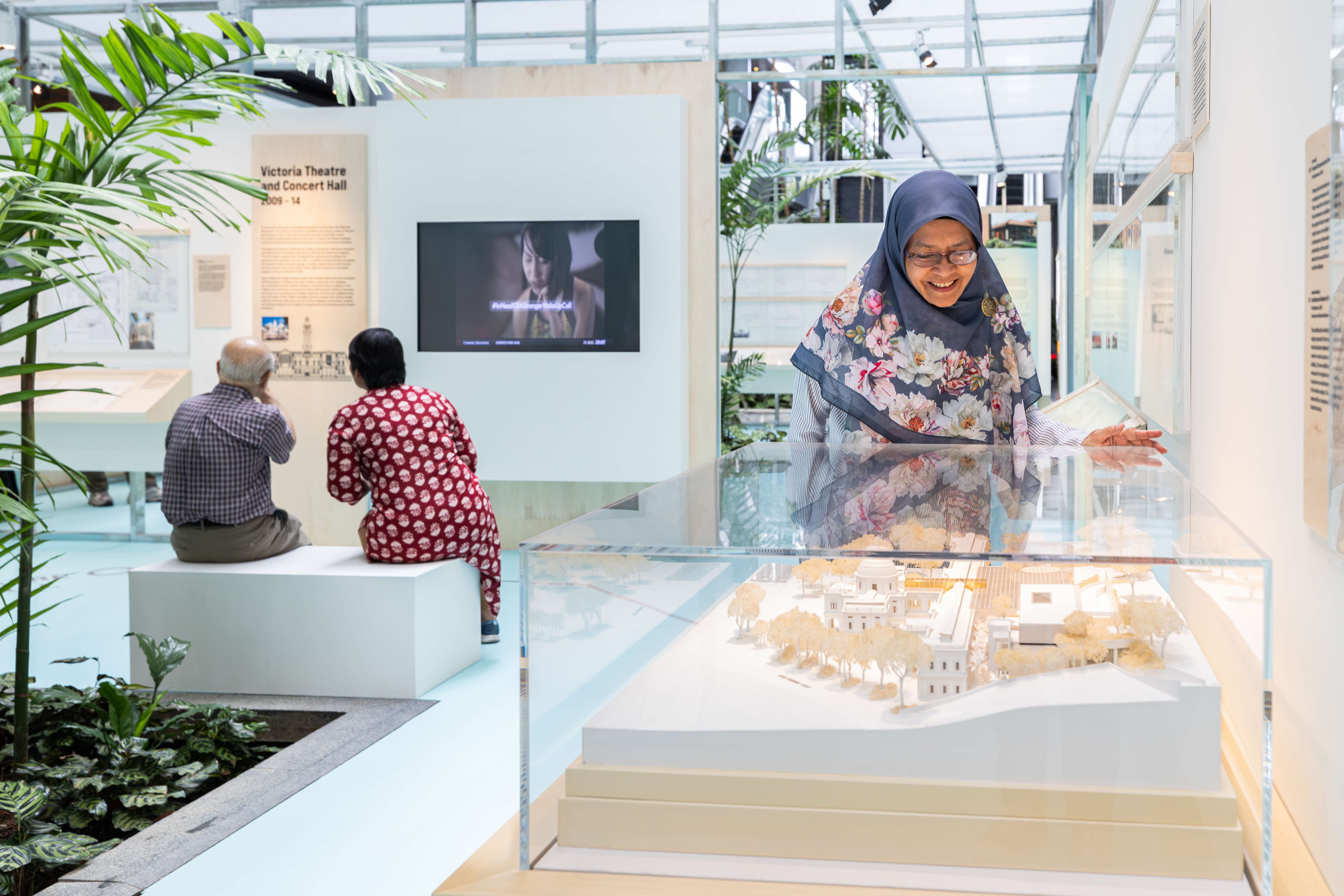 Two people are seated watching a screen, while a person inspects a miniature architectural model in an indoor setting.