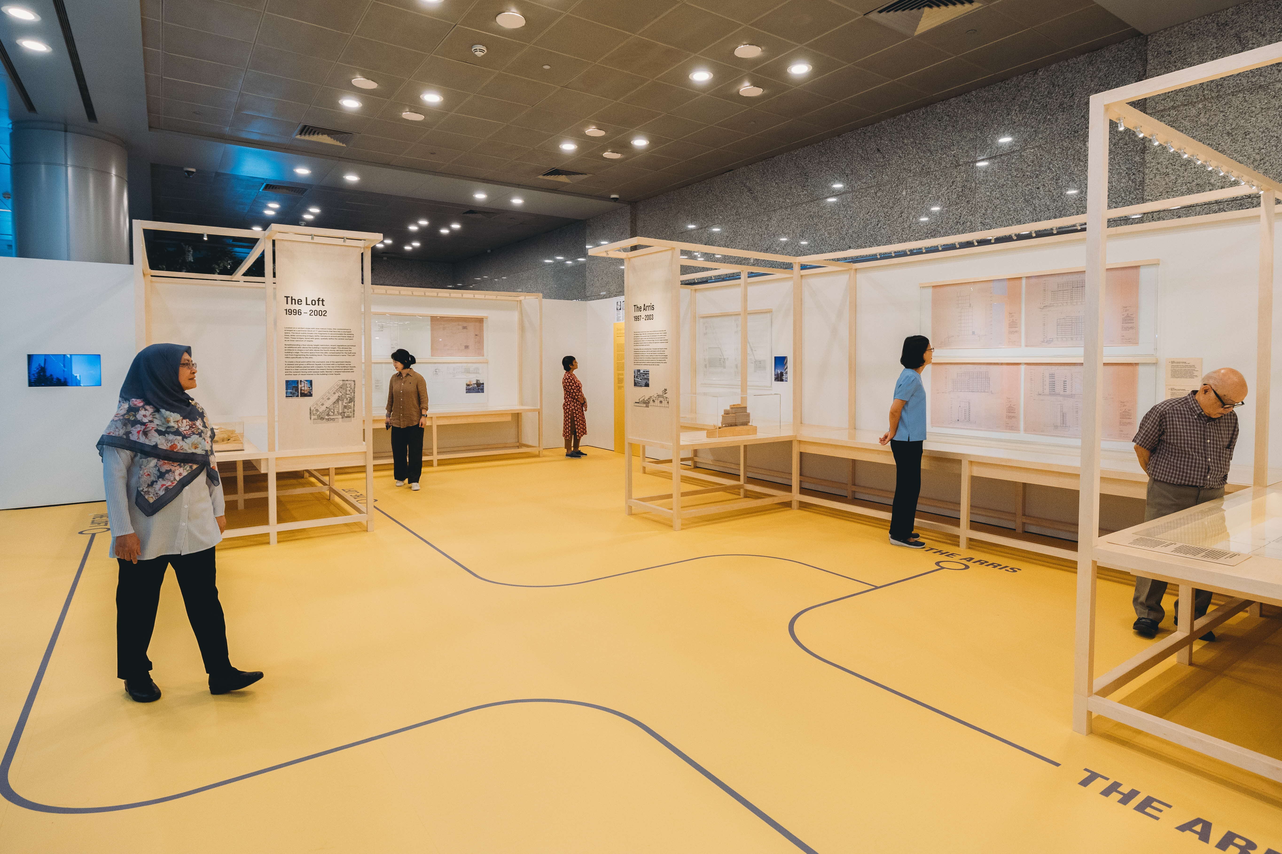 People are viewing architectural exhibits inside a brightly lit museum with wooden frames and yellow flooring.