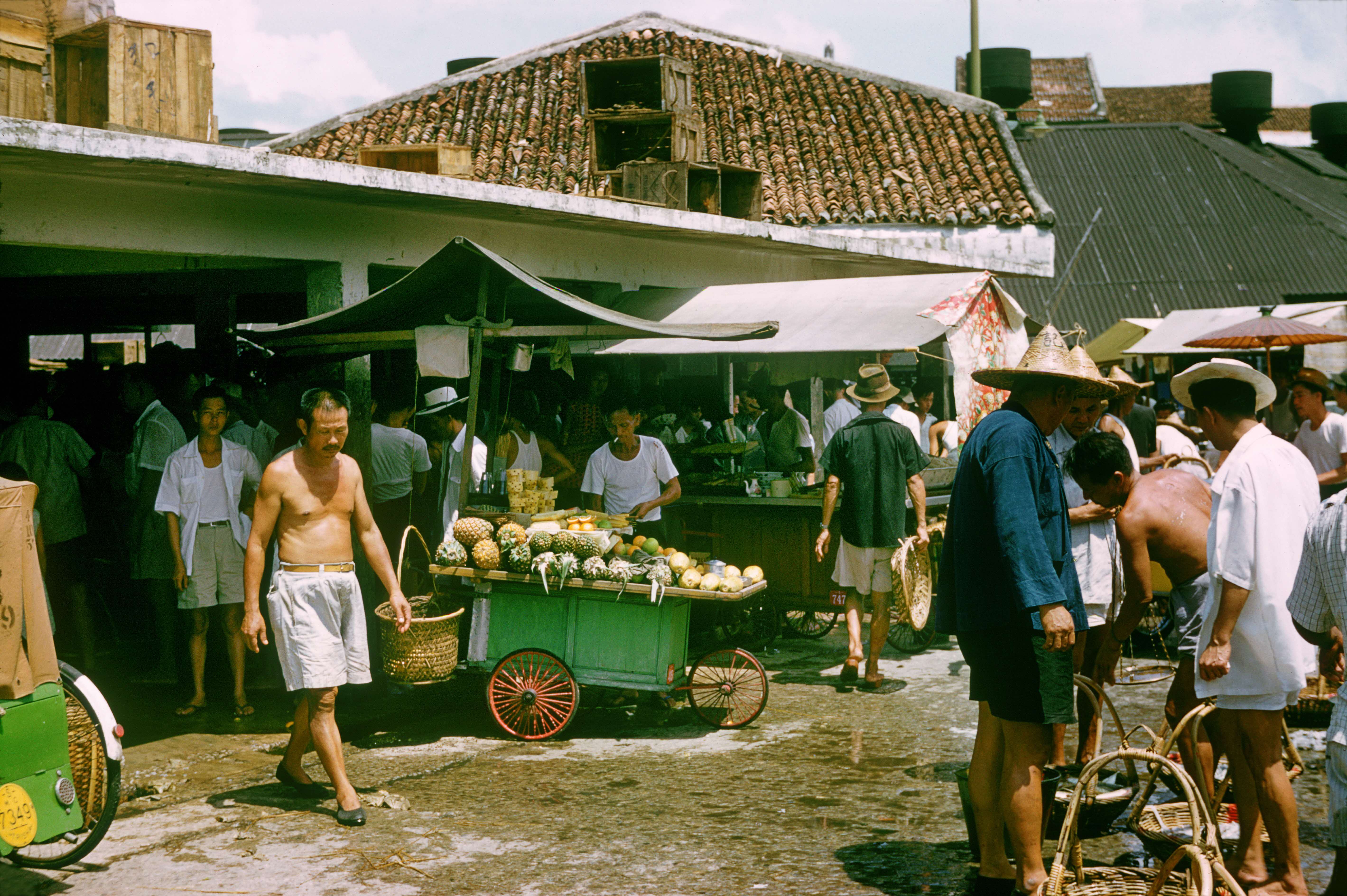 Outdoor market scene with people browsing fruit stalls under canopies, near a building with a tiled roof.
