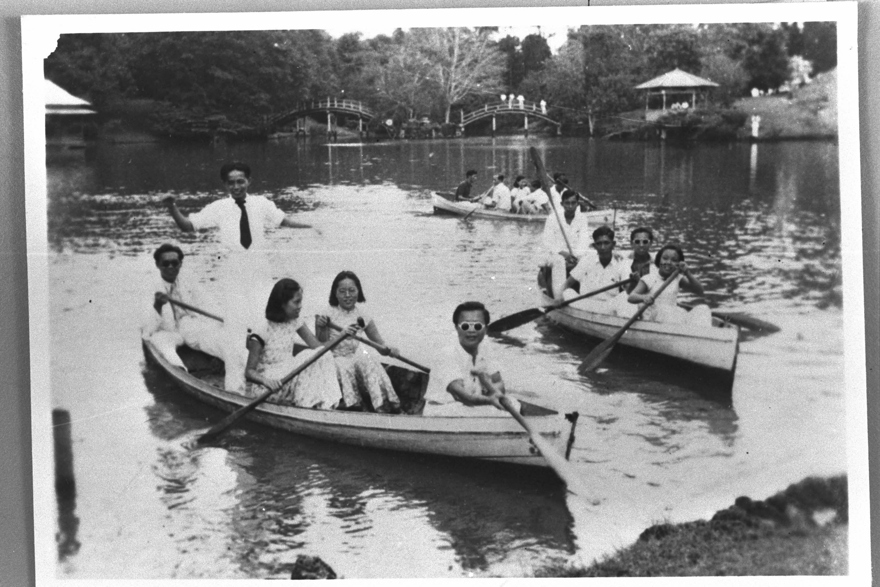 People row boats on a lake, with a bridge and gazebo visible in the background.