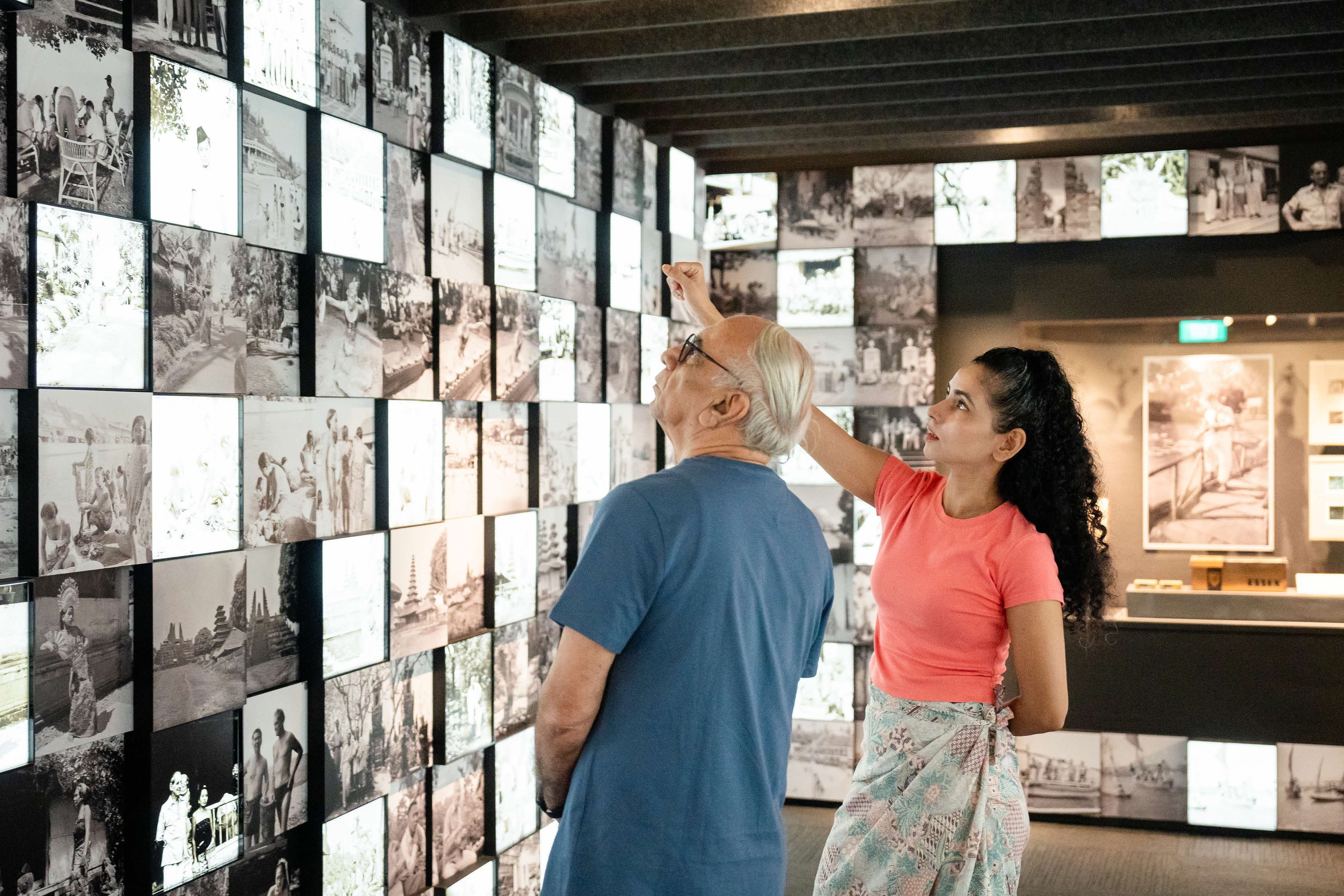 Two people stand in front of an illuminated wall full of black and white historical photographs in a museum.