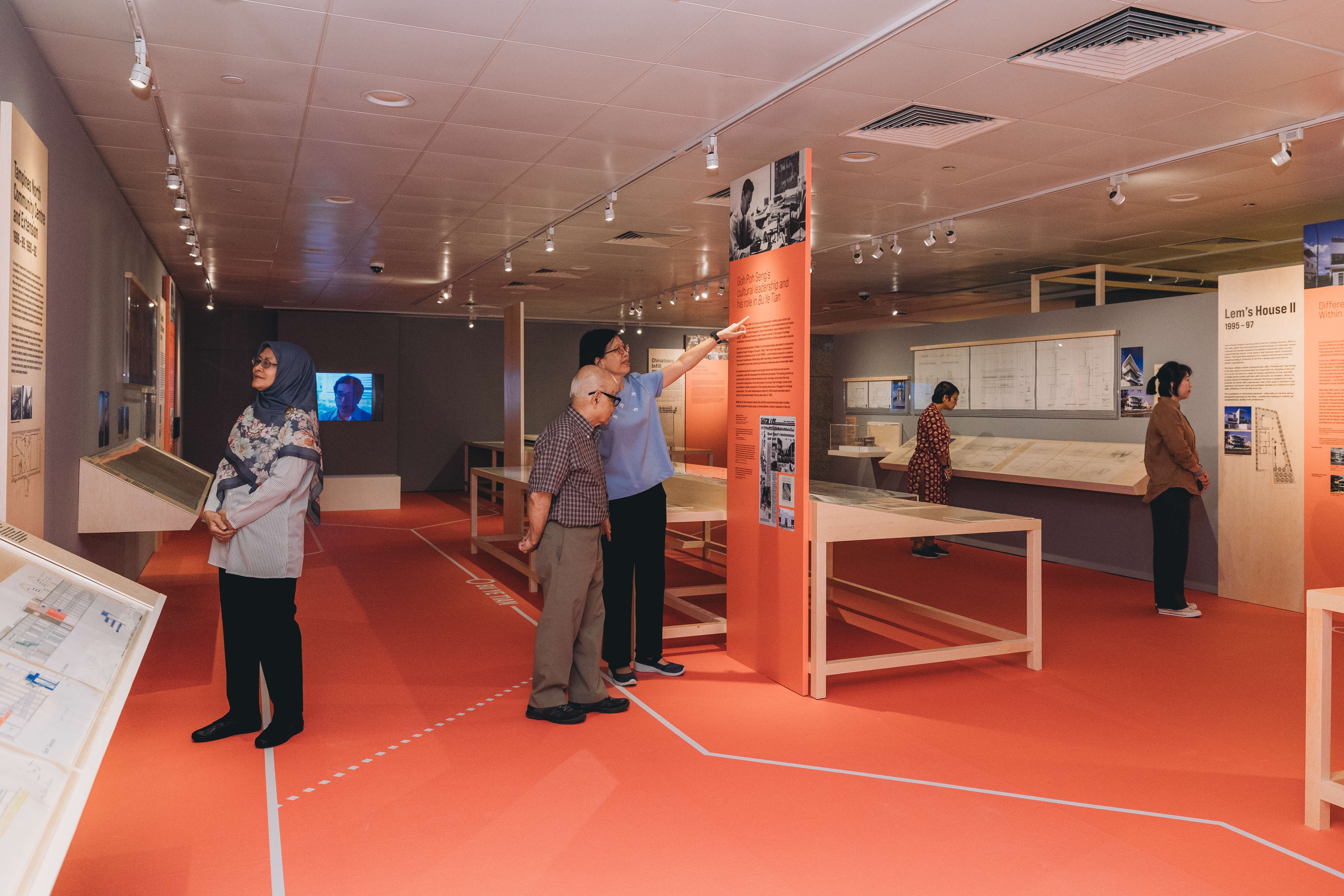 Four people observe displays in a brightly lit exhibit room with red flooring, informational panels, and table models.