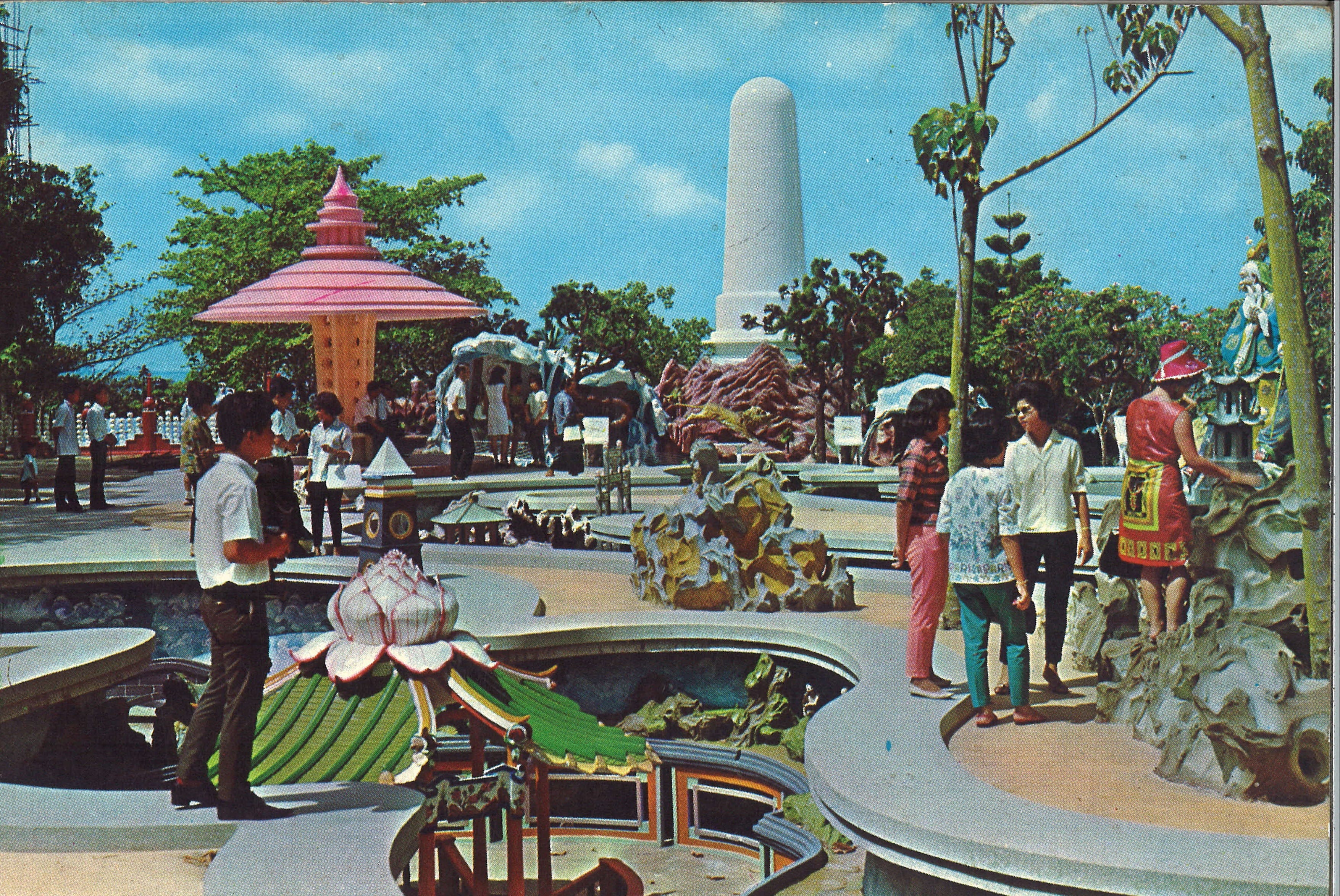 People are walking and sightseeing in an outdoor park with sculptures, greenery, and a large white pillar.