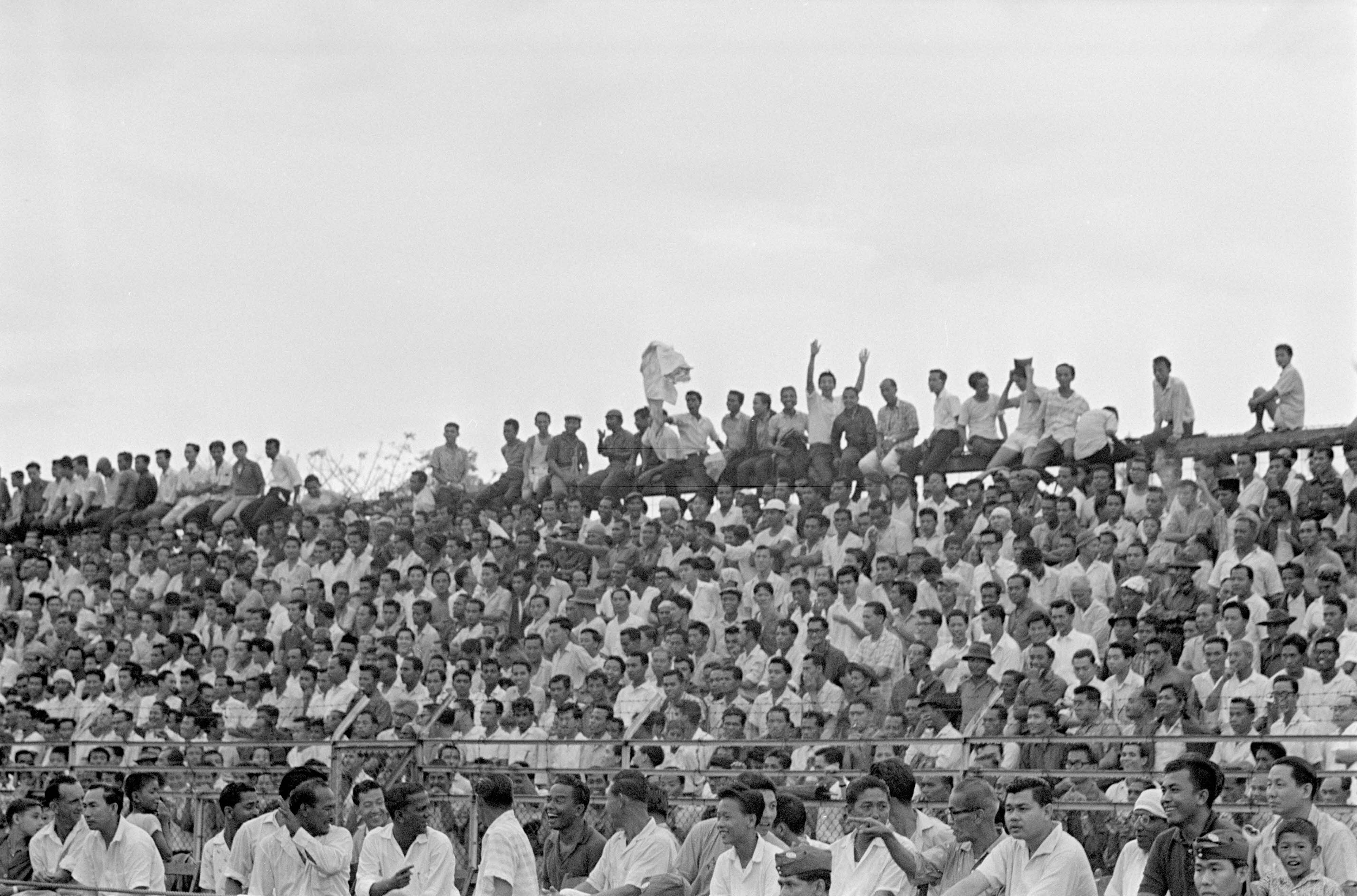 A large crowd of people, some sitting and others standing, gathered in an outdoor stadium or event area.