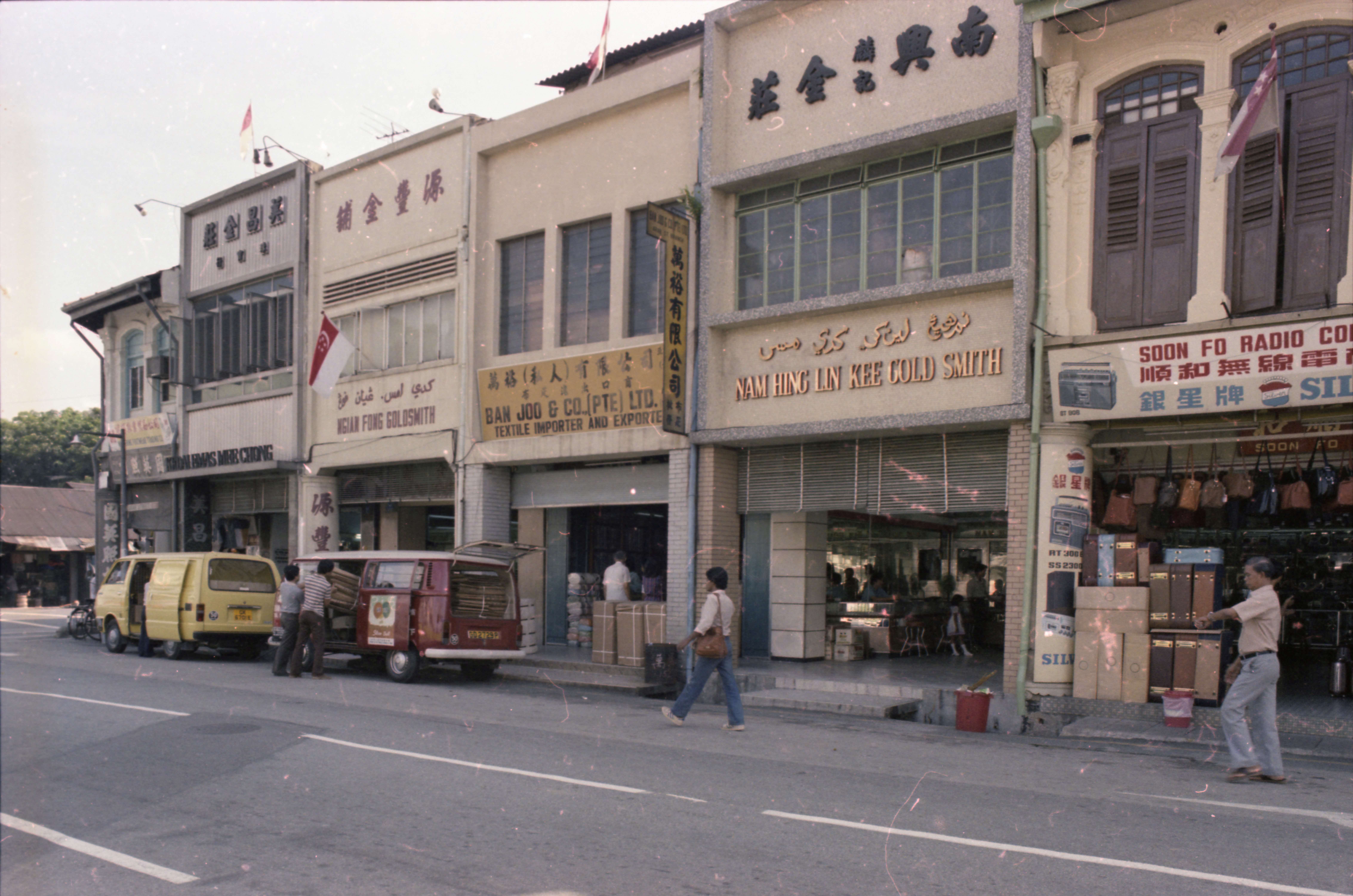 A row of shops, including a goldsmith, textile importer, and radio company with vehicles parked and people walking by.