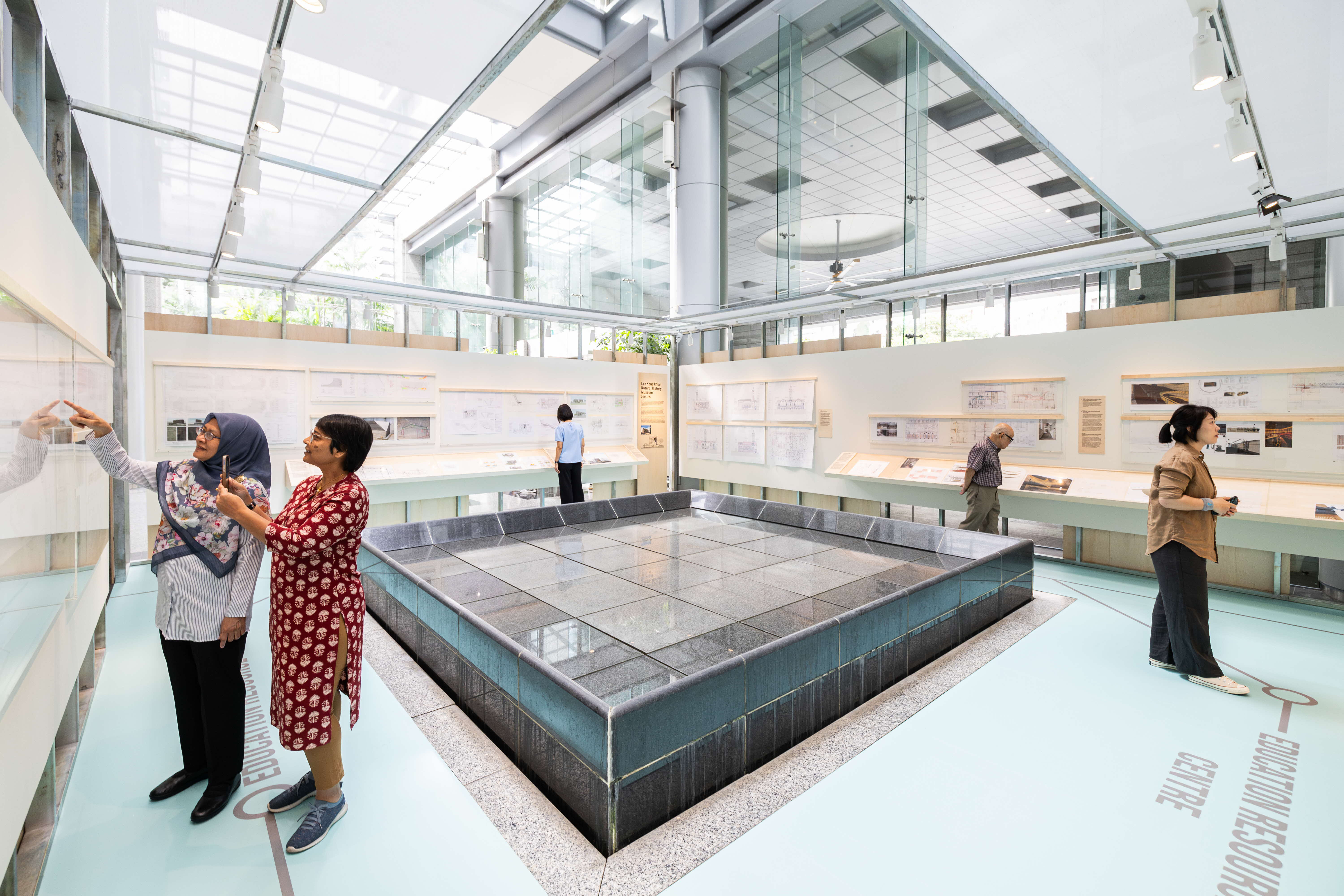 A modern exhibition room with large windows, diagrams on the walls, and five people examining the displays.