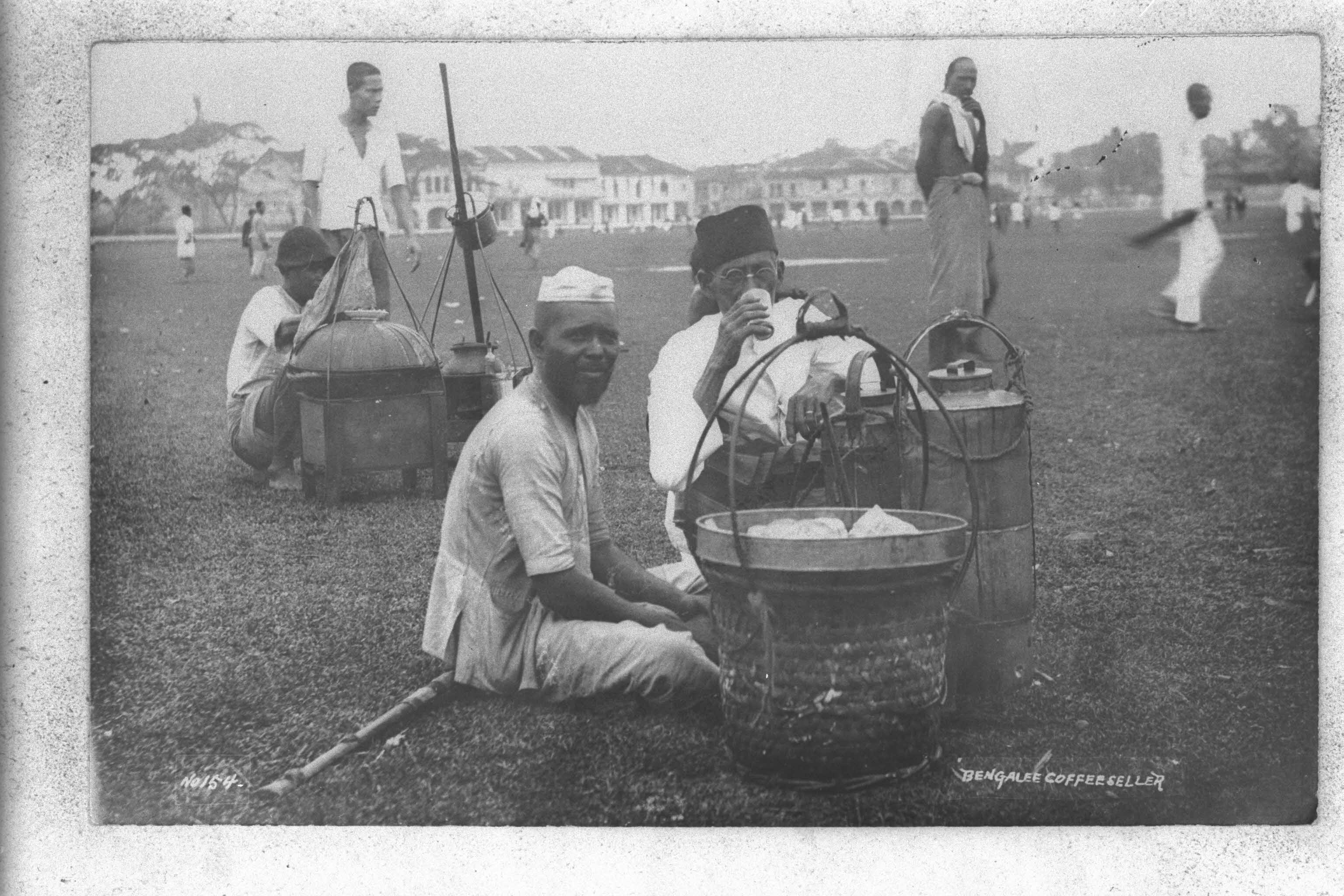 A group of men sit on grass with containers, with buildings and additional people in the background.