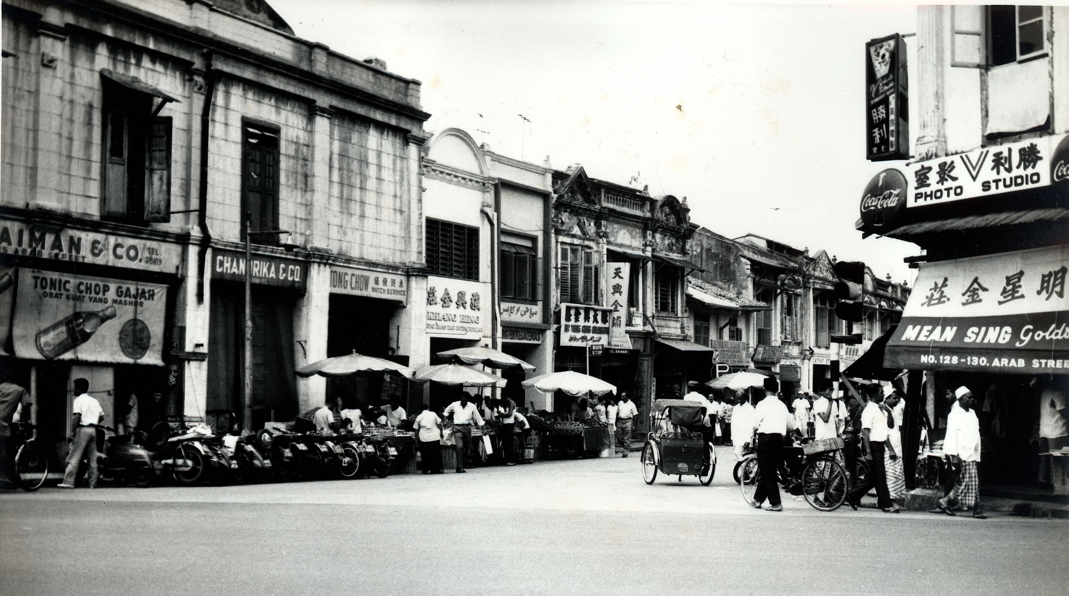 Street scene with several people walking and shopping near a row of old buildings housing various shops.
